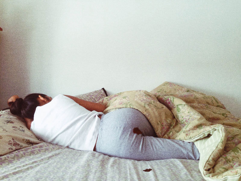 Woman on her period bleeding through her trousers, lying on her bed