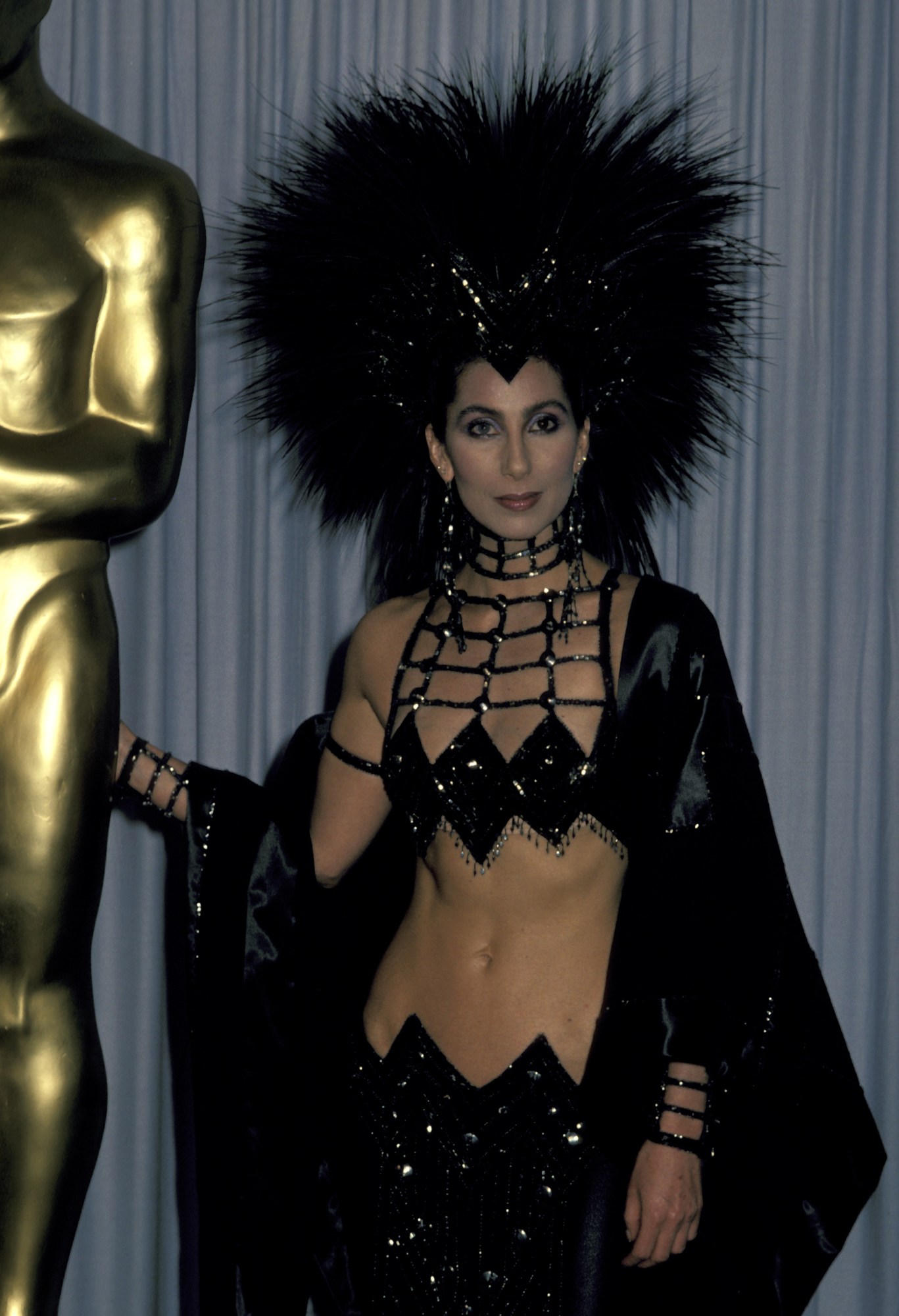 cher posing at the academy awards in a two piece sequin dress and feather headress by bob mackie