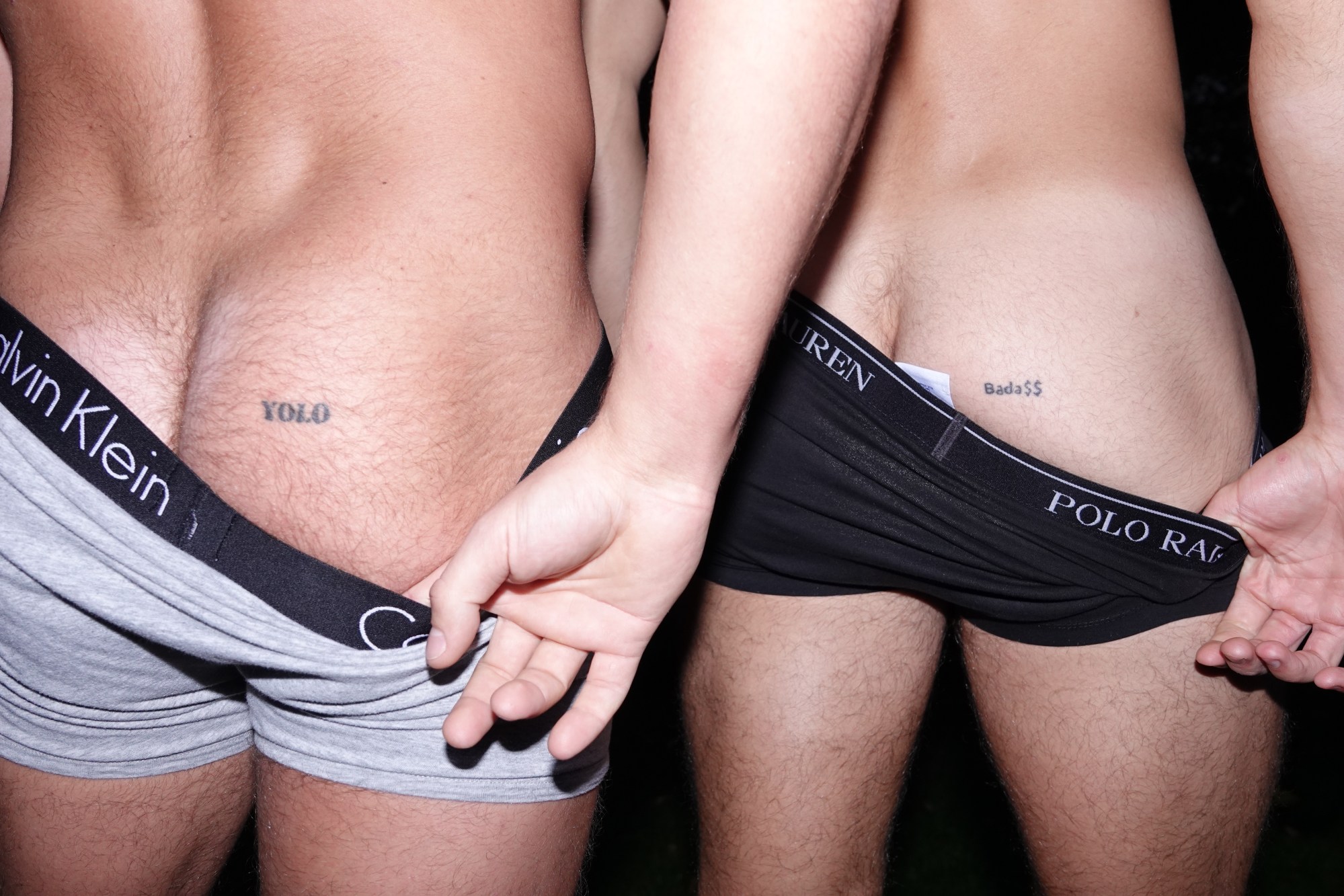 An image of two men, each pulling down the other's boxers to reveal a tattooed buttock.