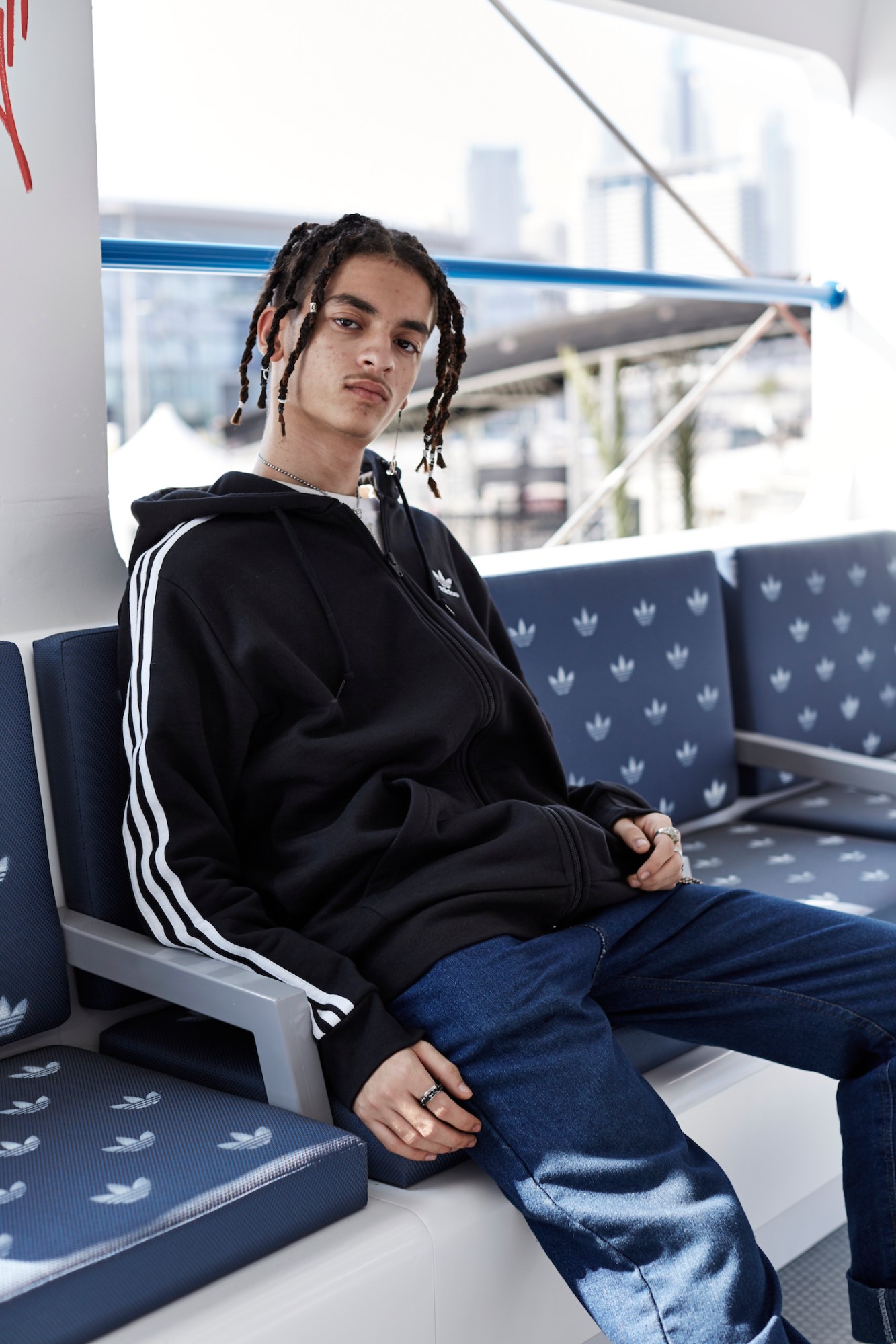 A young man with dreadlocks wear a three-stripe Adidas jacket and sits on an Adidas logo-ed metro seat.