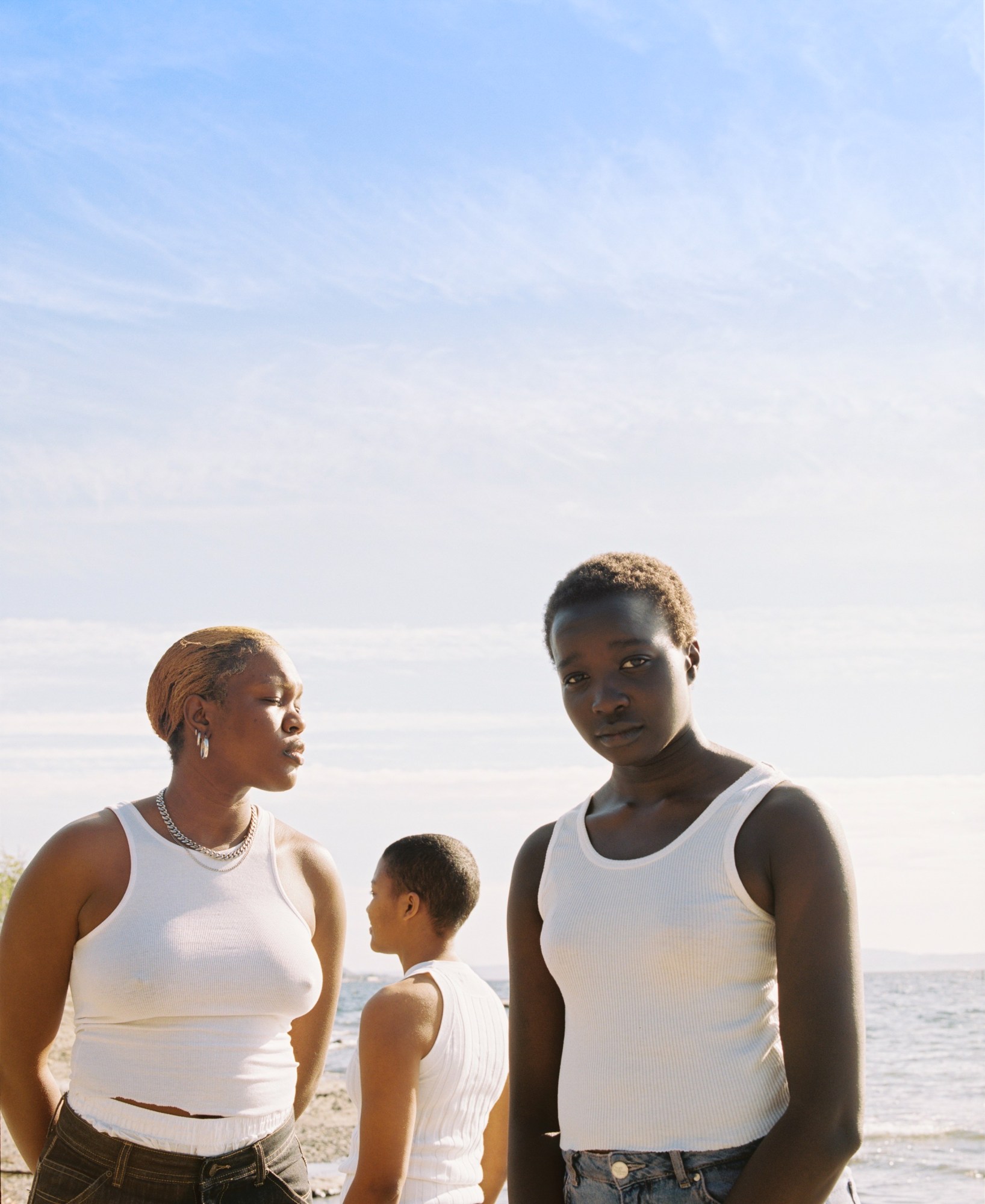 three people stood on the beach in the sun, each wearing white vest tops