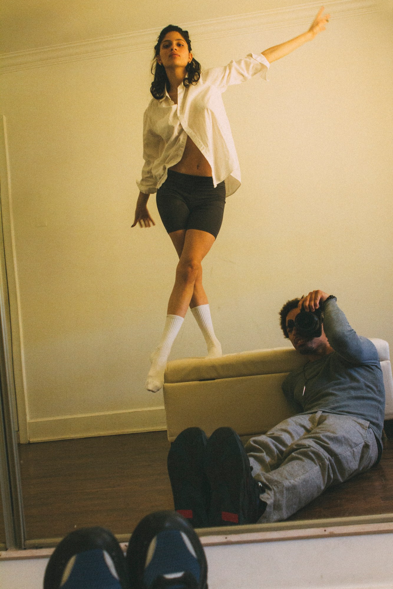 a man sits on the floor, reflected back in the mirror, taking a photo of a woman in a shirt and lycra shorts standing above him