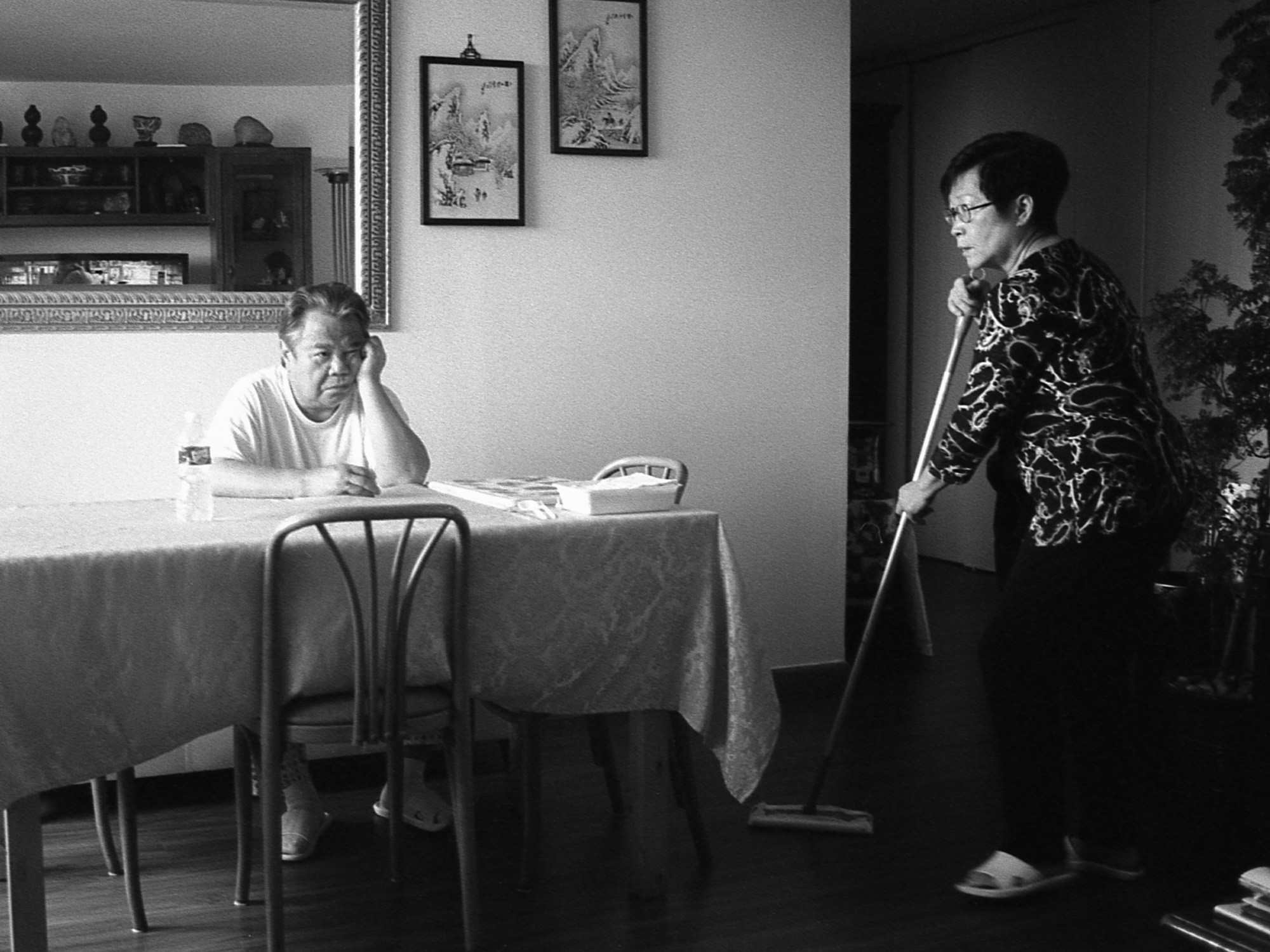 a photo of an man sat at a table with his head resting on his hand and a woman sweeping the floor next to him