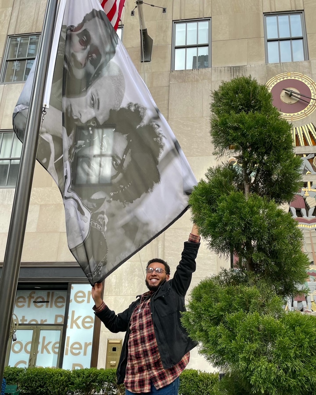 a young man in a plaid shirt, black jacket and blue jeans holds a flag with one of their own photographs on it flying outside the rockefeller centre