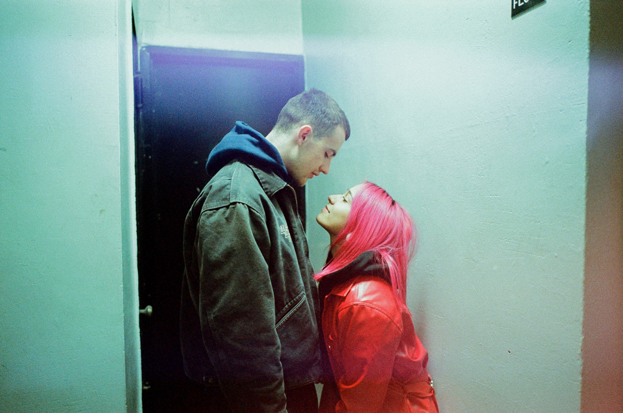 a tall man with short hair looks down at a woman with pink hair and a bright red coat in an apartment hallway