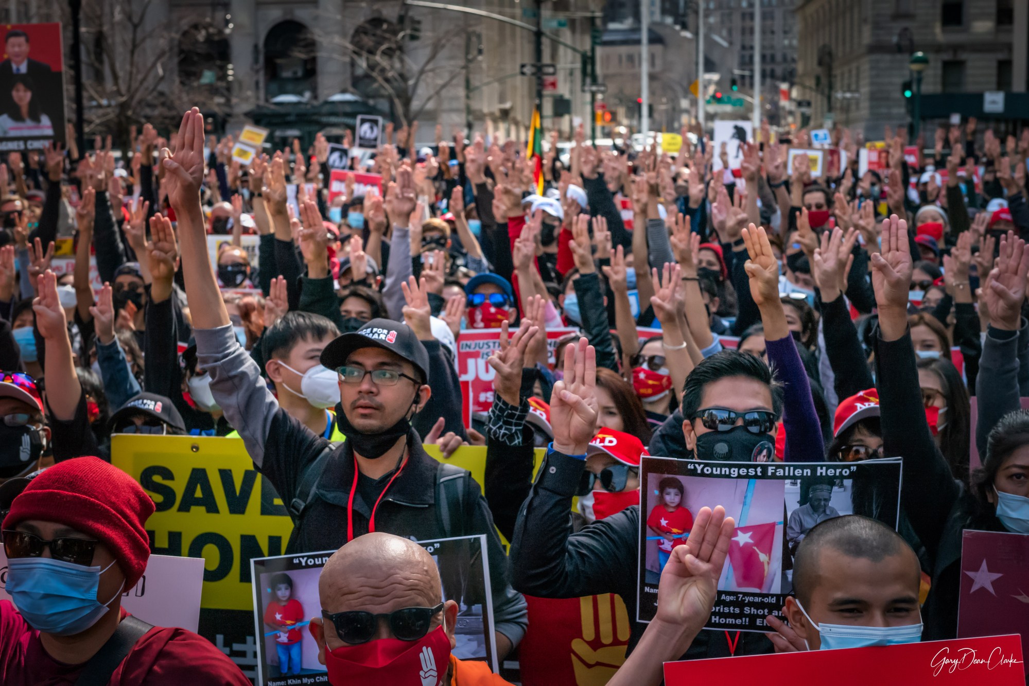 a large crowd of protestors, many of whom are wearing red, hold up three fingers