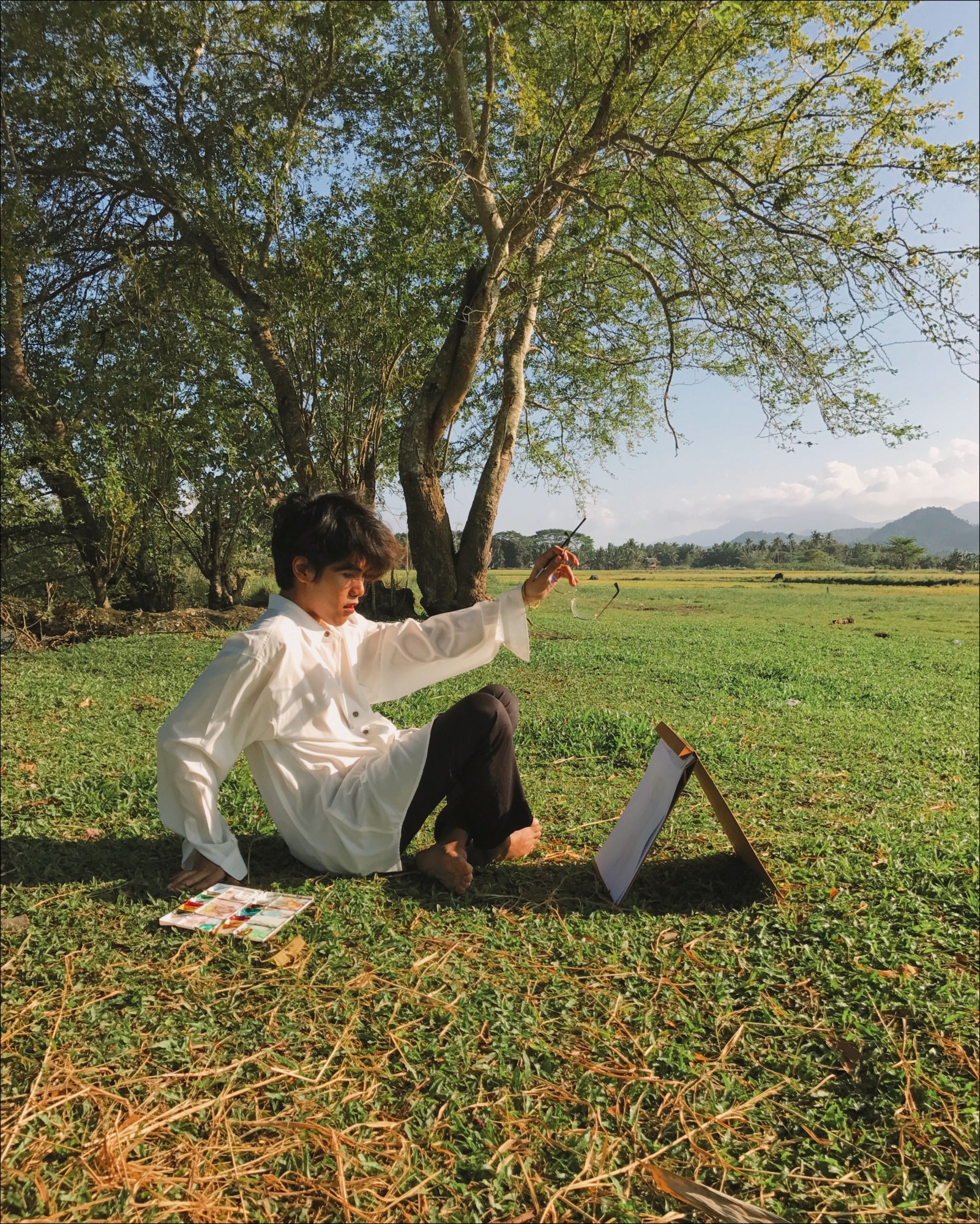 a man in a long white shirt sits in a field painting