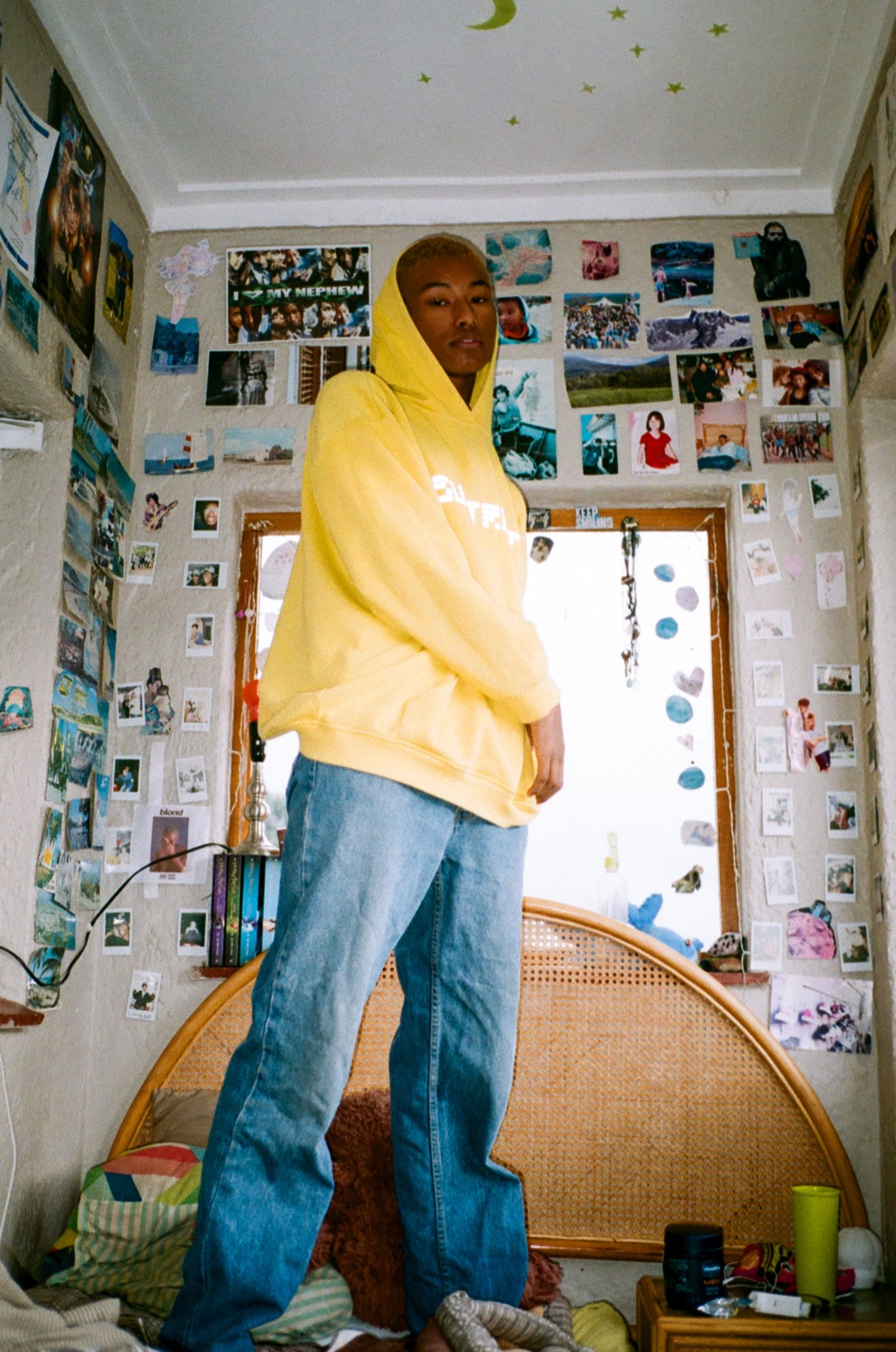 a man in blue jeans and a yellow hoodie stands on his bed in front of a wall covered in pictures