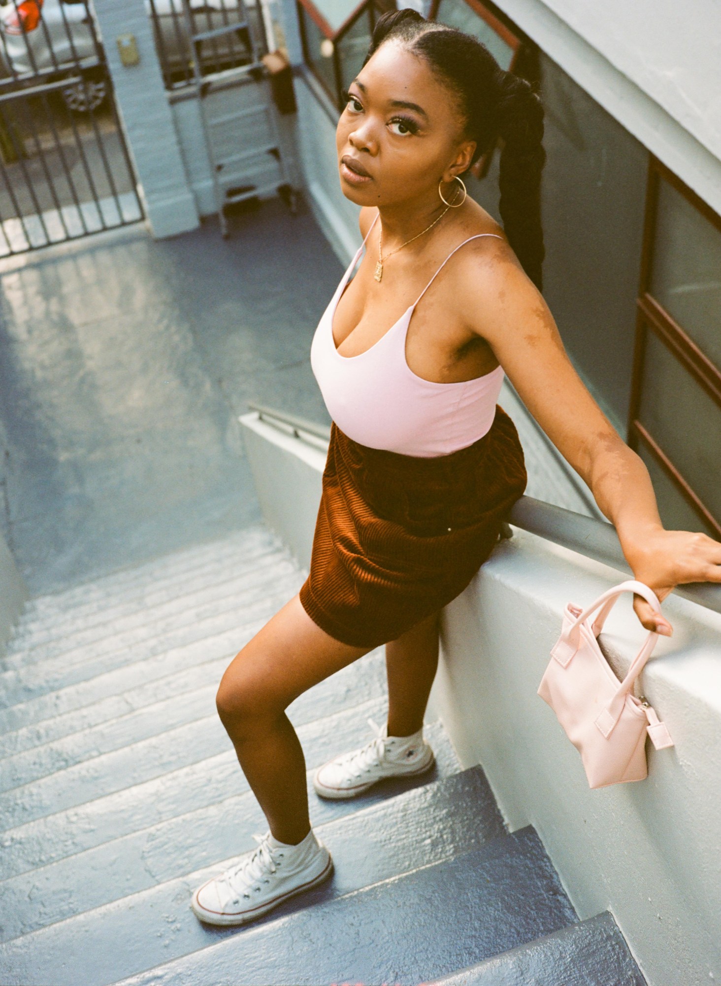 a woman standing on a staircase wearing white converse, a brown corduroy skirt and a pink top