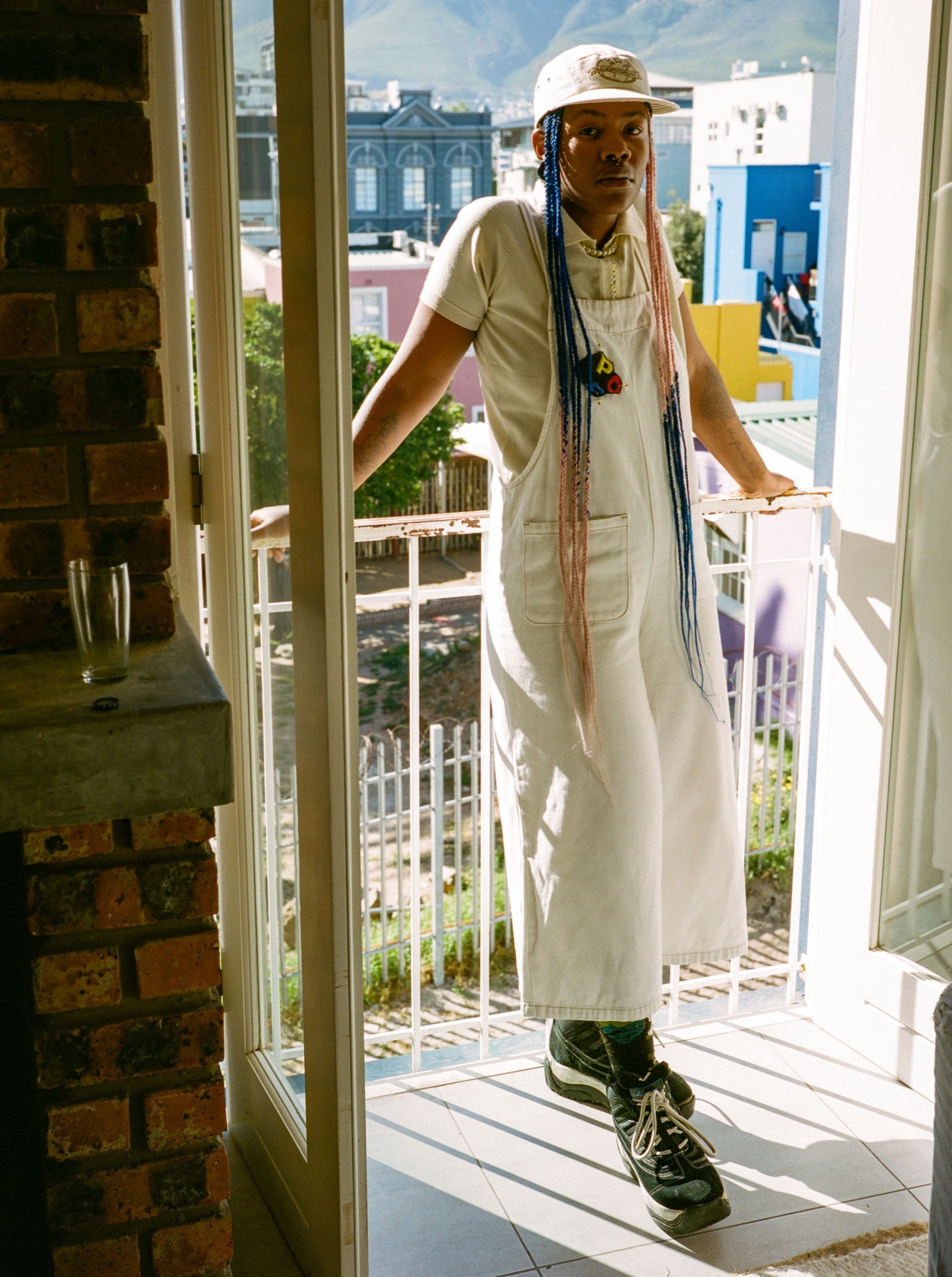 a woman in white dungarees holds onto the railings outside an apartment window