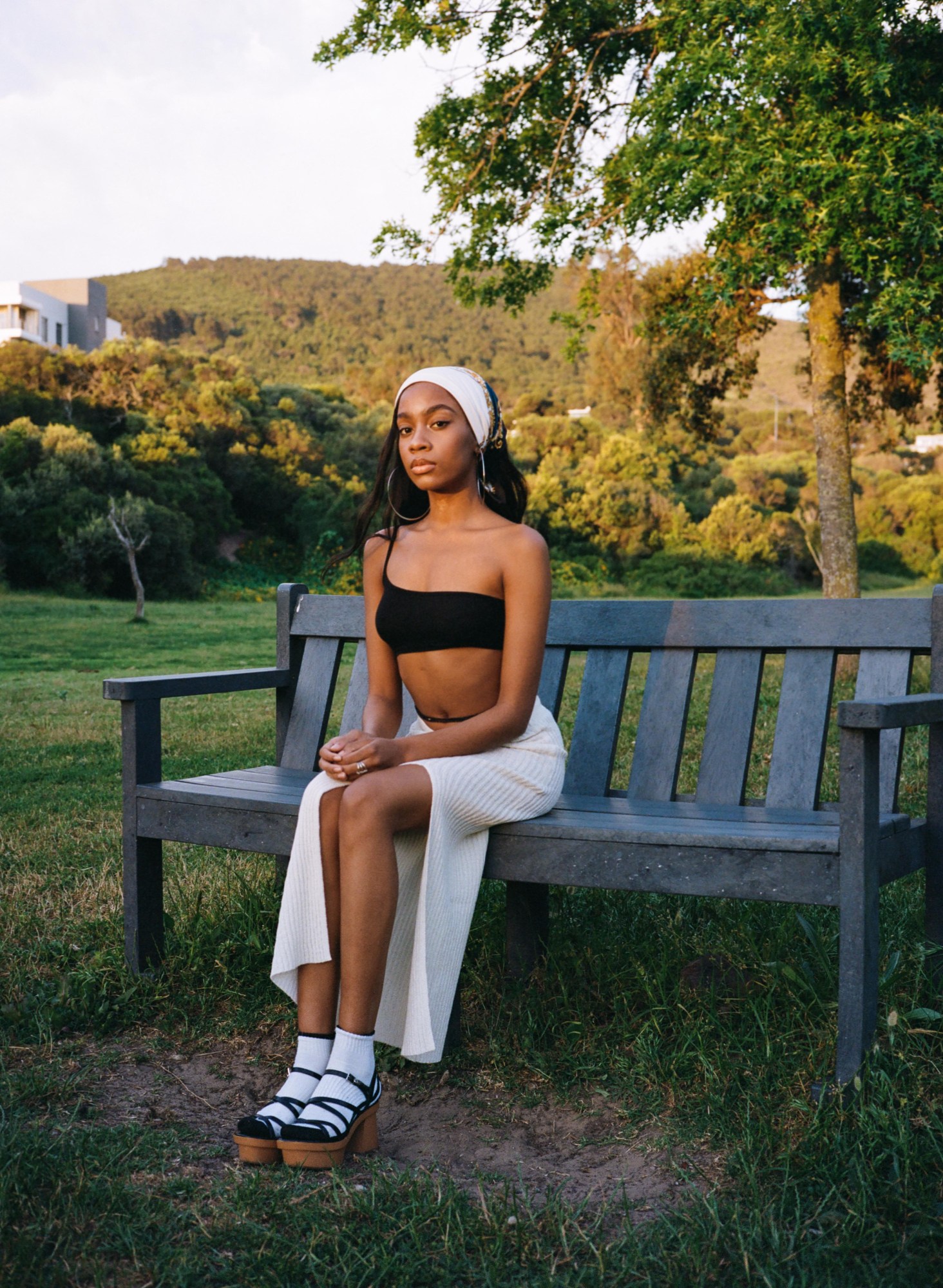 a woman sits on a bench in a park wearing a long white skirt and heels