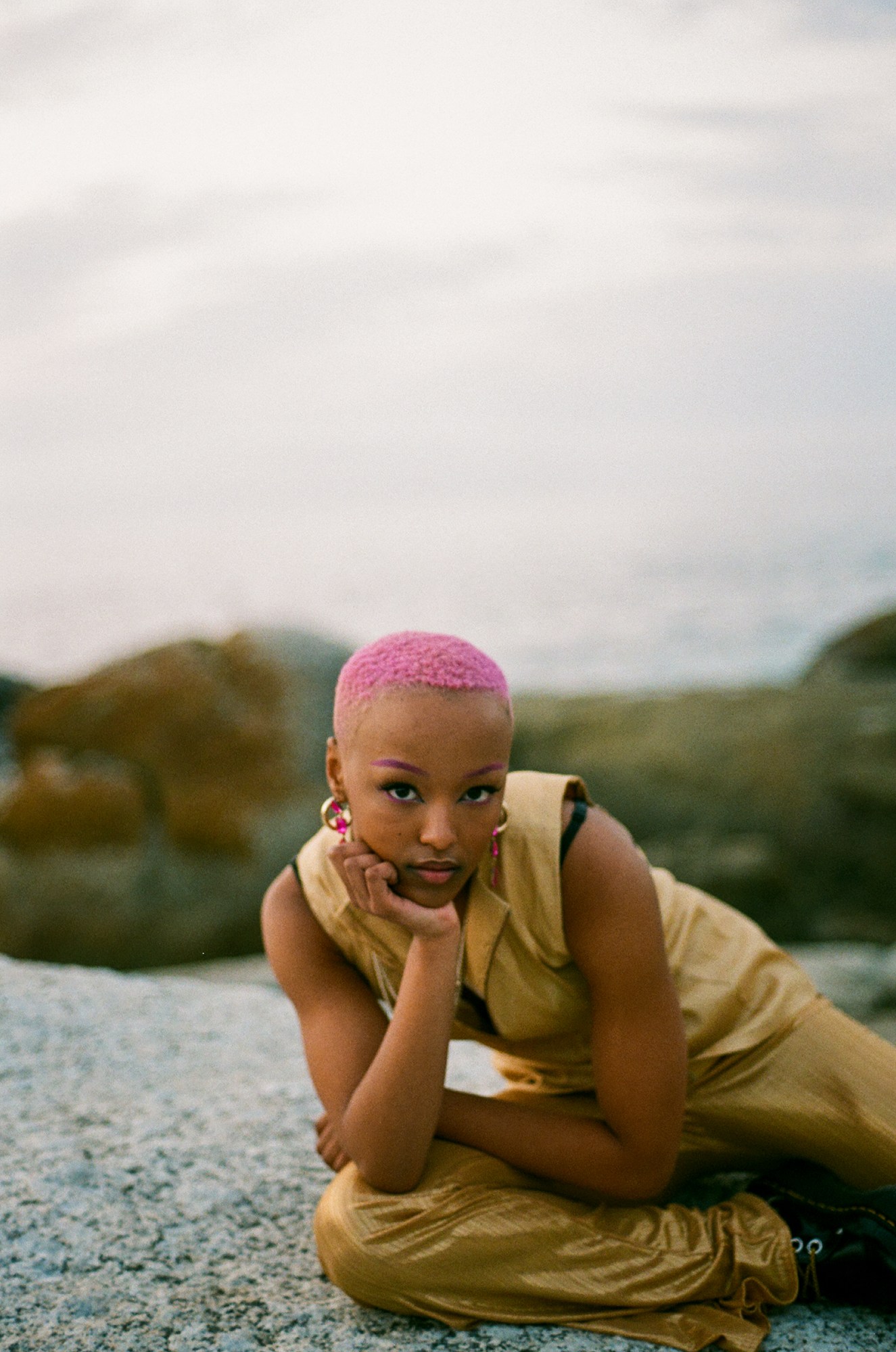 a woman with pink hair and pink eyebrows wearing a gold jumpsuit