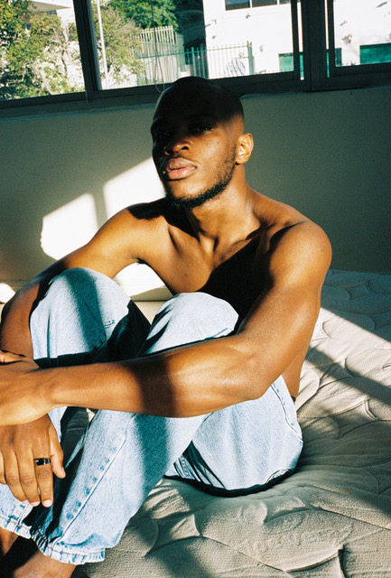 a topless man wearing blue jeans sits on a mattress with a shadow covering half his face