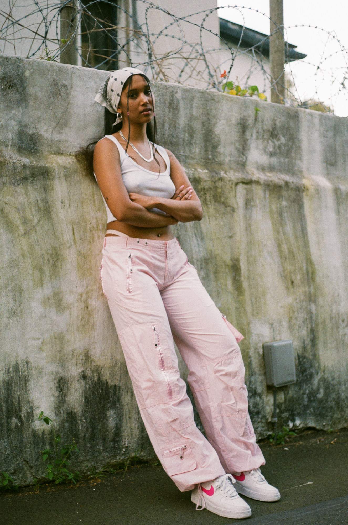 a woman with a headscarf, white top and baby pink trousers leans against a wall with her arms crossed