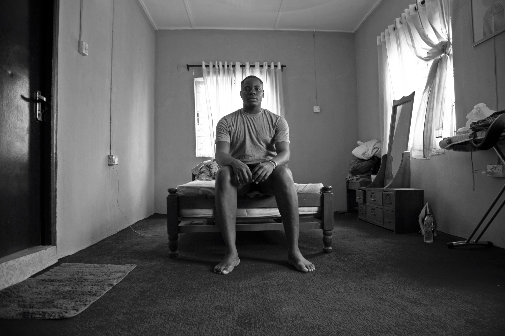 a man sits at the end of a bed in a bedroom with two windows