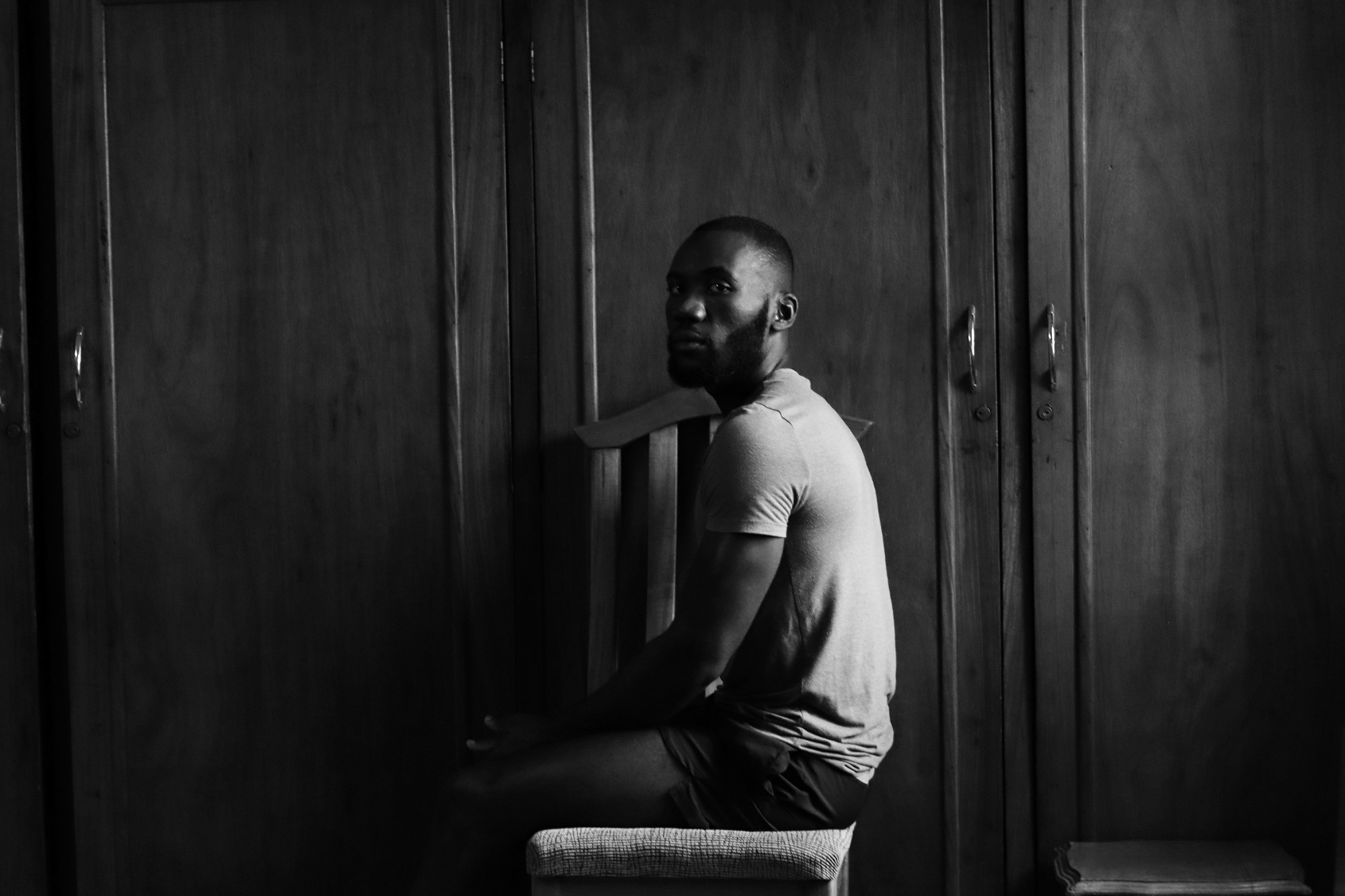 a man sits sideways on, looking over his shoulder at the camera, in front of cupboards