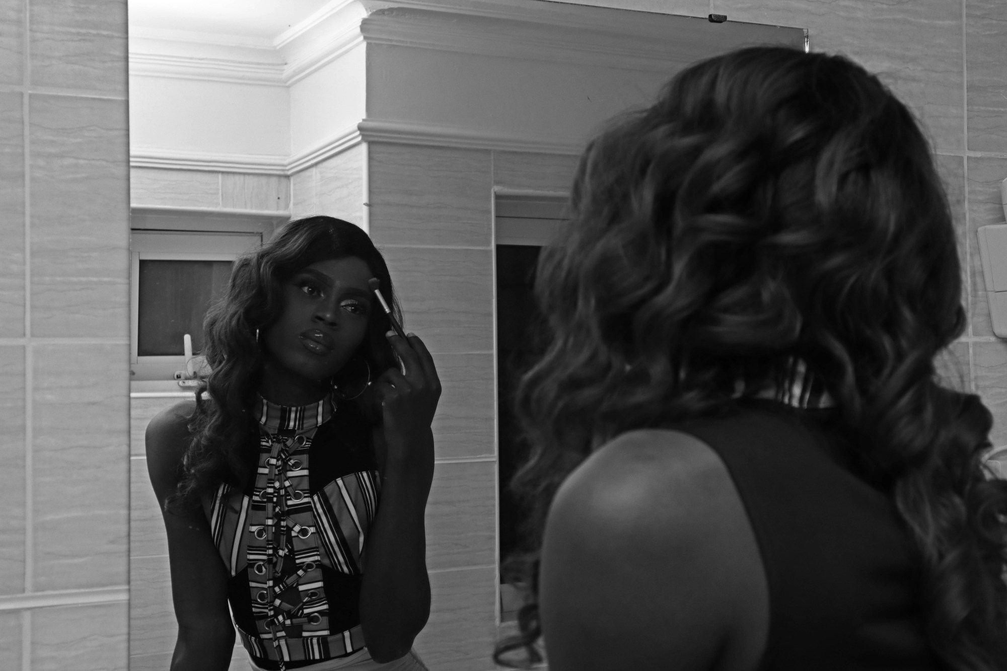 a person in makeup and a wig looks into the mirror holding a makeup brush