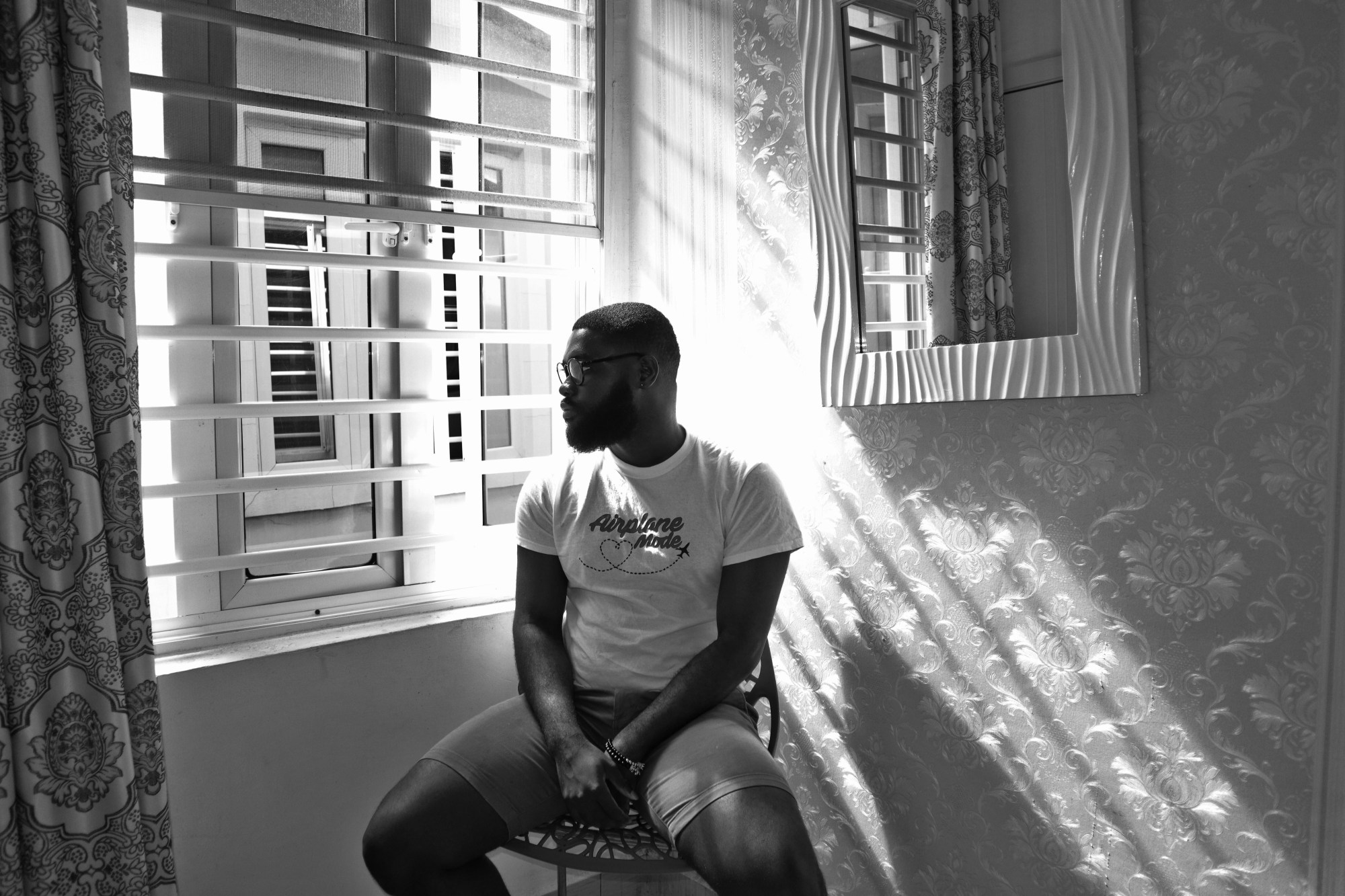 a man in a white t-shirt and tight shorts sits in the corner of a room next to a window with half open blinds