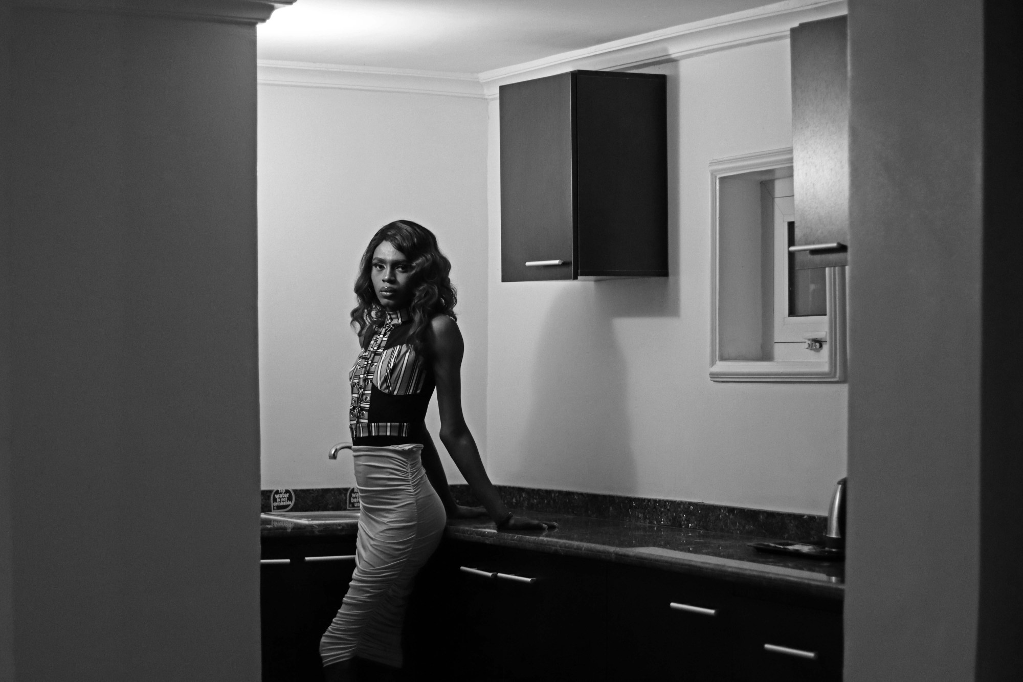 a person stands in the corner of a kitchen dressed in a tight dress and wig