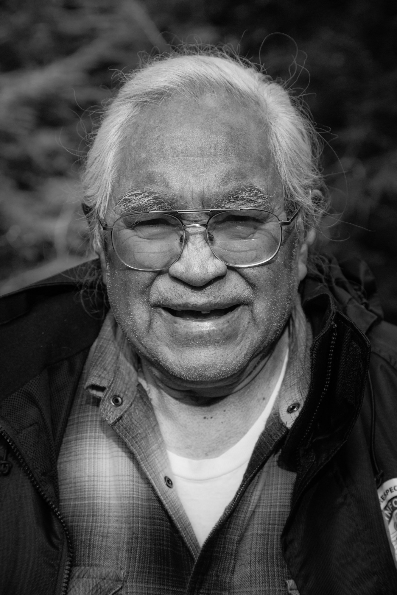 an old white-haired Pacheedaht man wearing clear aviator glasses and a plaid shirt, looking to camera