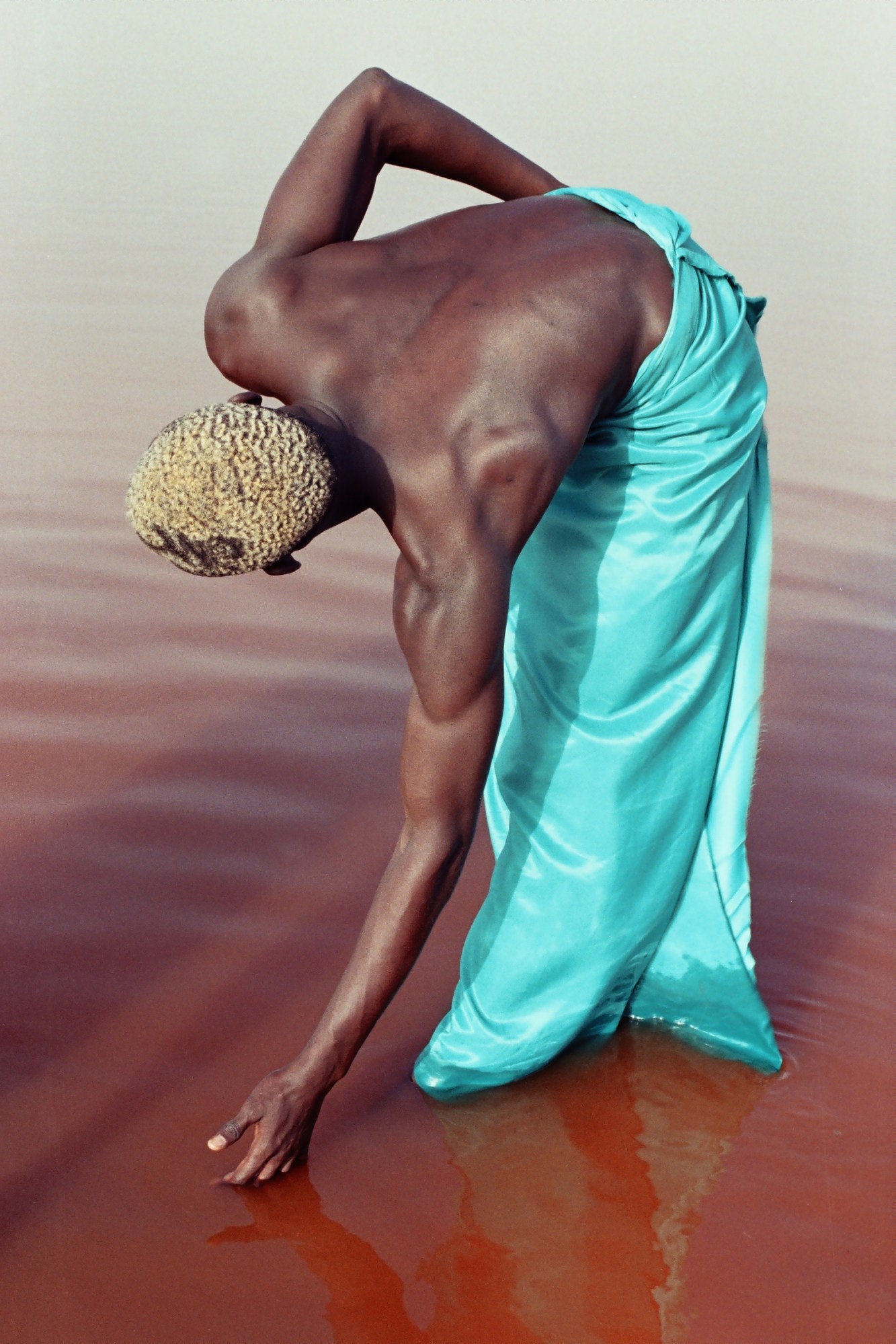 a man with bleached blonde hair and a turquoise robe lowers his hand into dark orange water