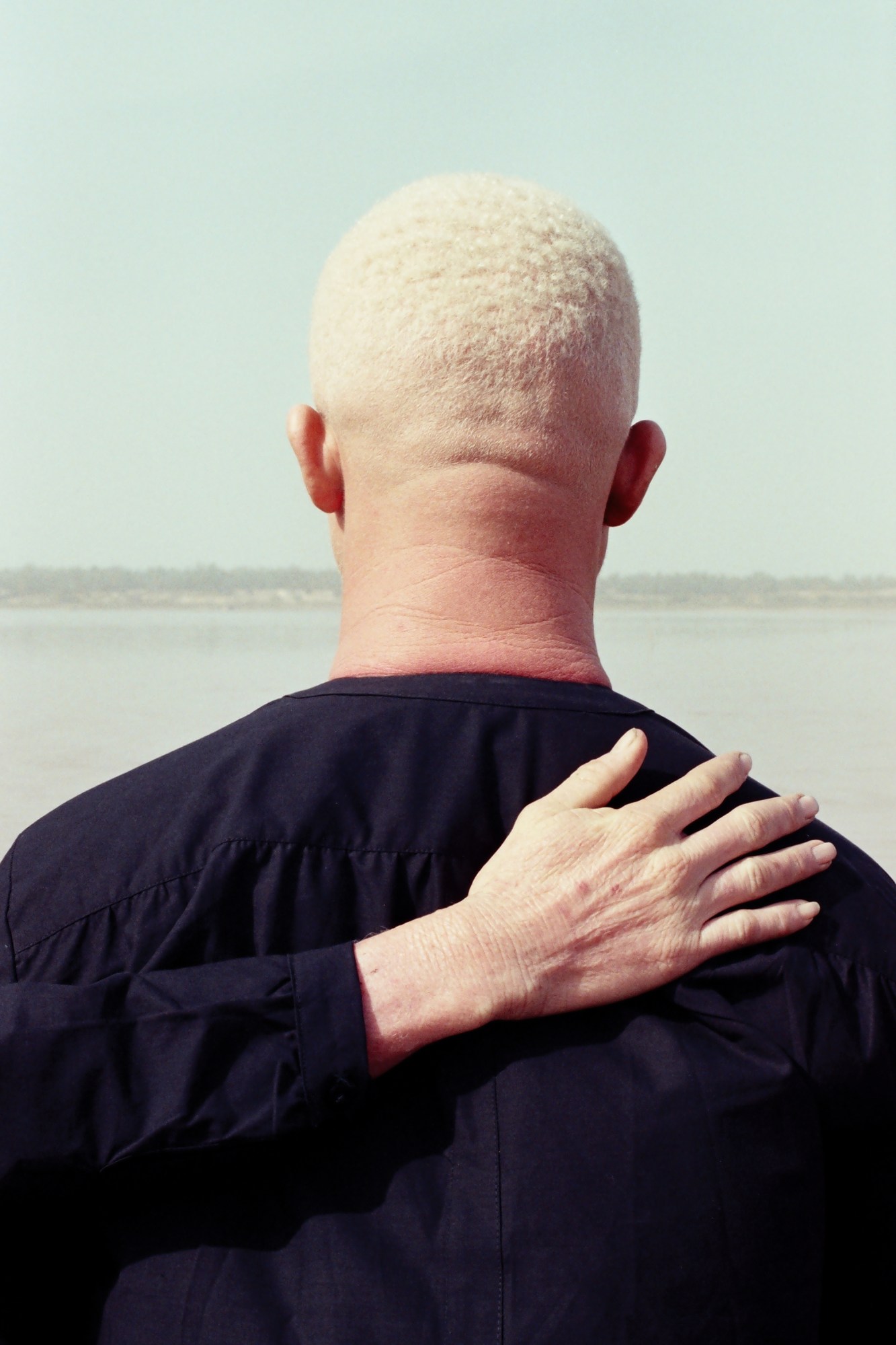 the back of a man's head and a hand resting on his shoulder looks towards a body of water