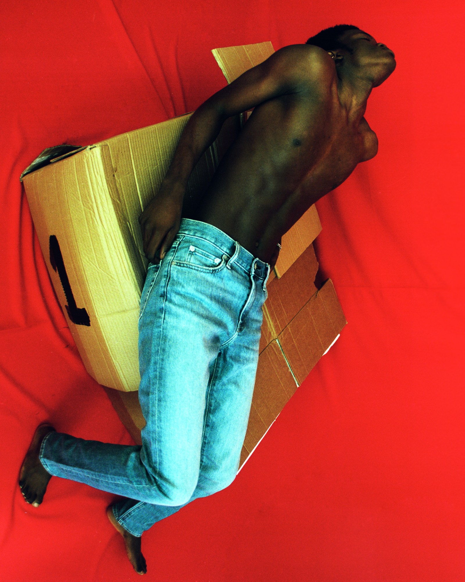 a man wearing no top and blue jeans sits on a cardboard box against a red backdrop