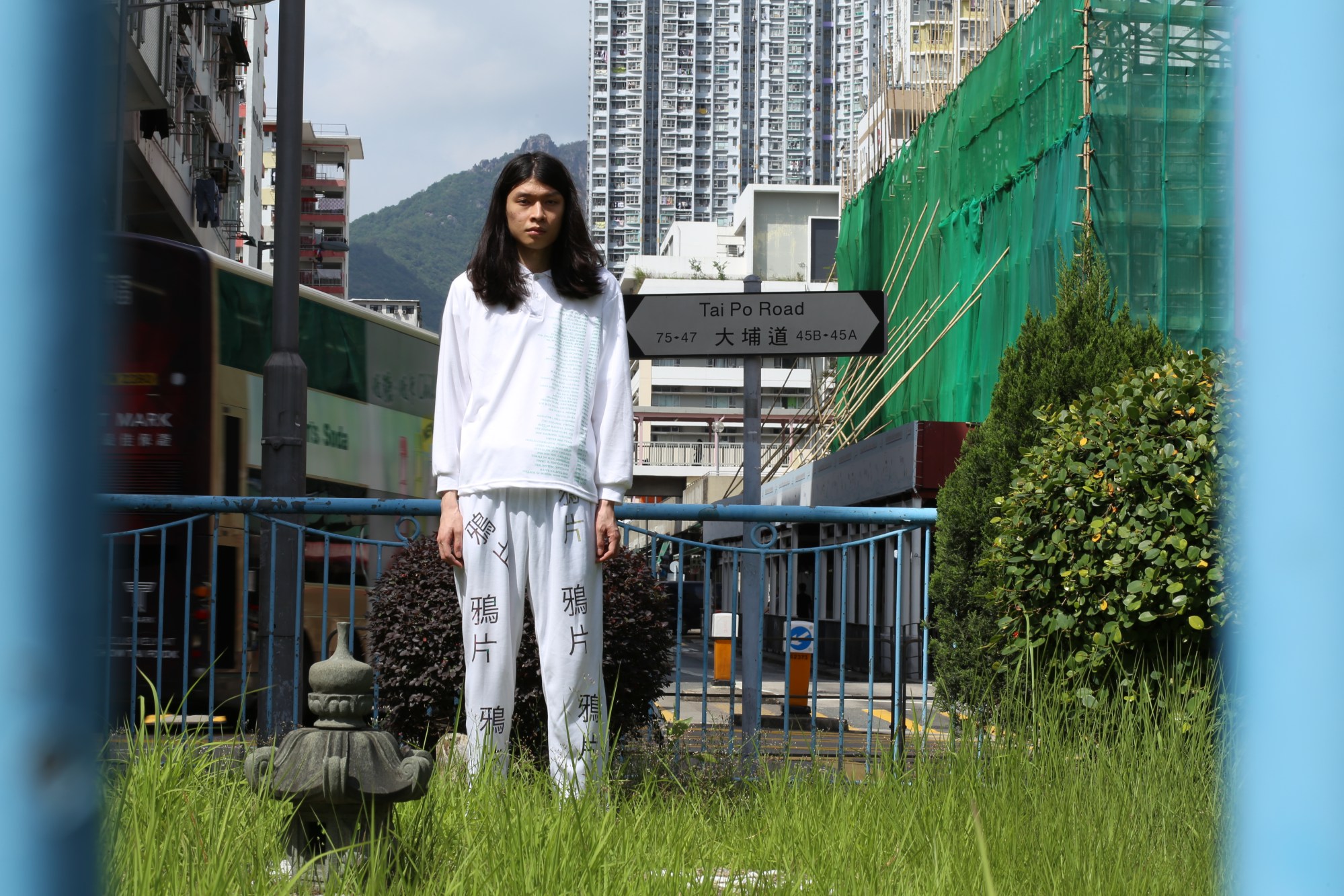 A model wearing a longsleeve white top and white trousers standing on a grassy verge with a Hong Kong street as background