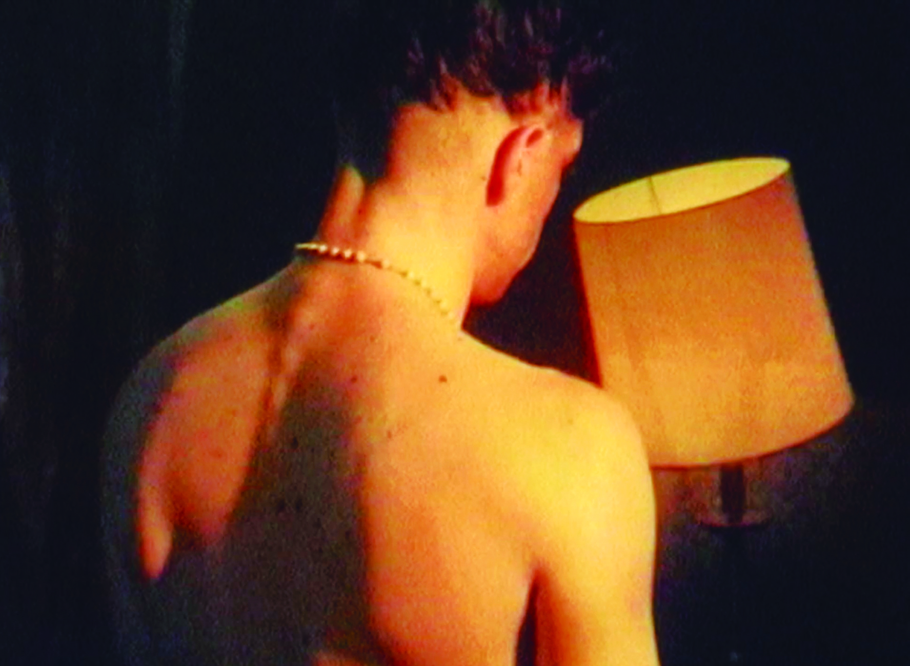 a topless man wearing a necklace, facing towards a lamp in front of him