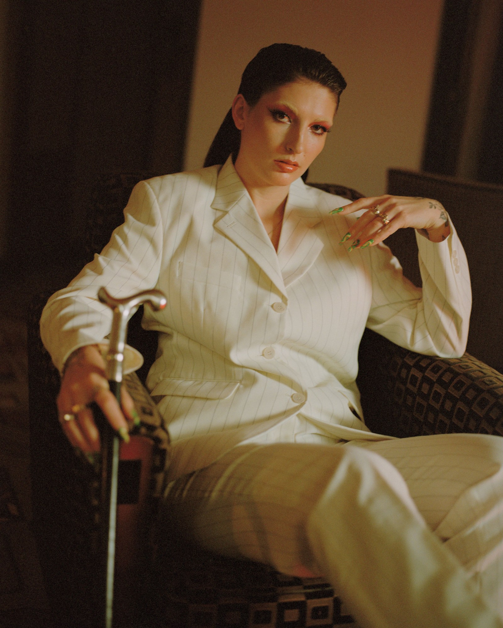 mila v wearing a white pinstriped suit, sitting in a patterned chair holding a cane