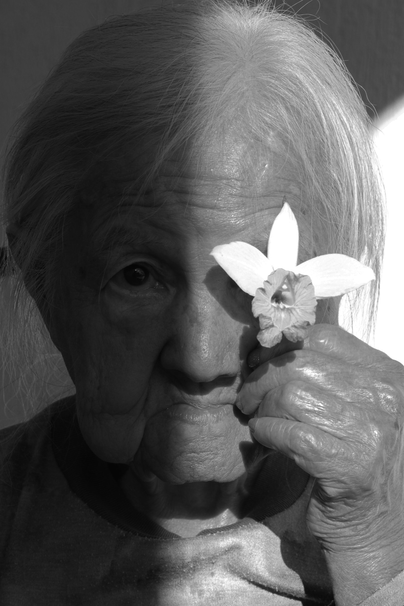 A black-and-white photo of a white-haired lady holding a flower to her eye.