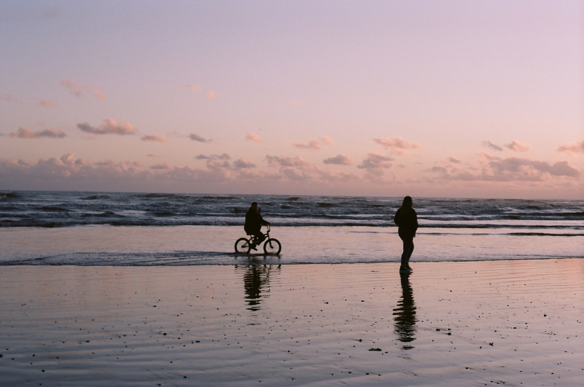 Two people on a beach at low-tide at dusk. One rides a bike, the other walks.