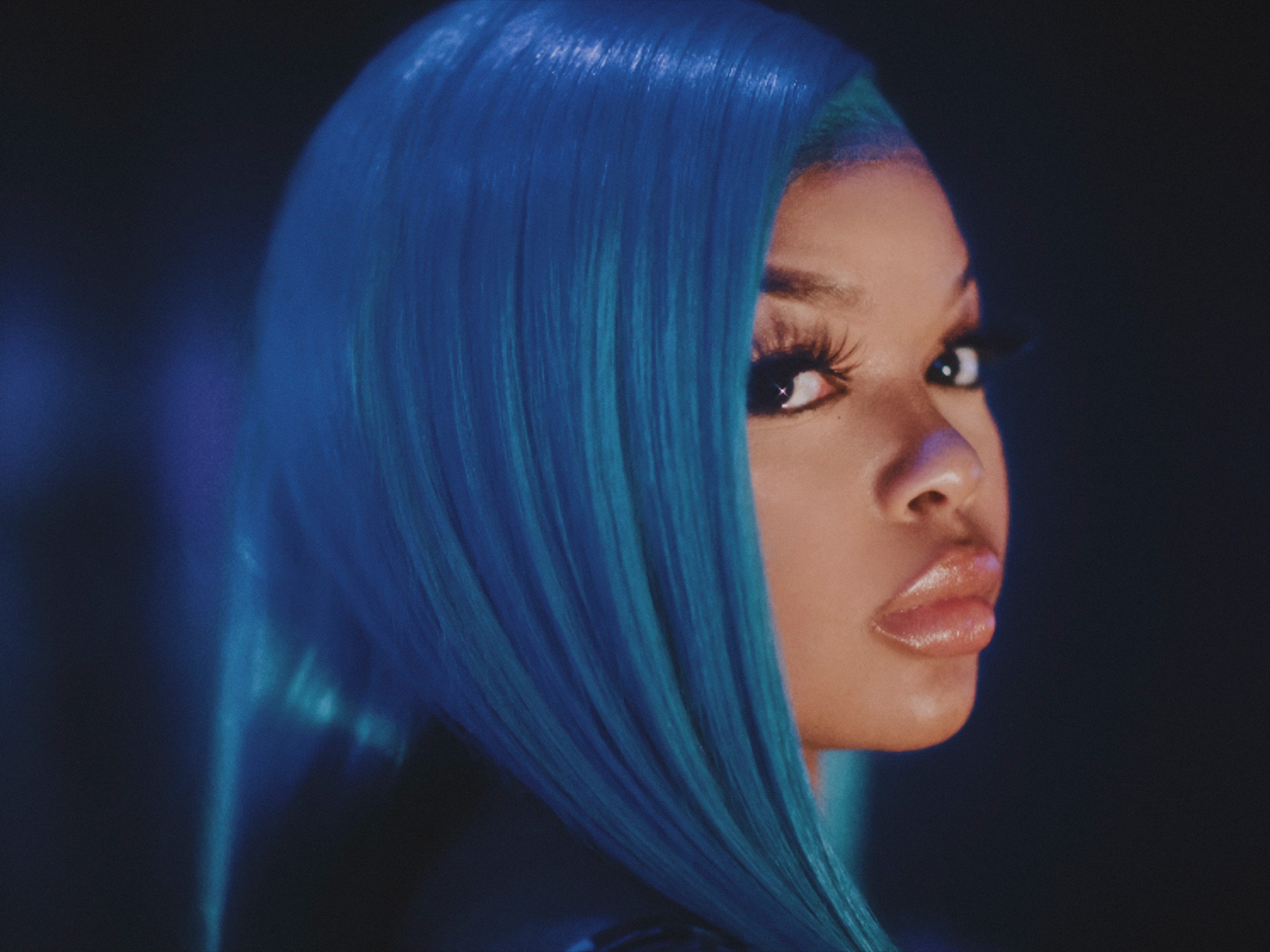 a woman with bright blue hair, fake lashes and glossy lips looks over her shoulder to camera