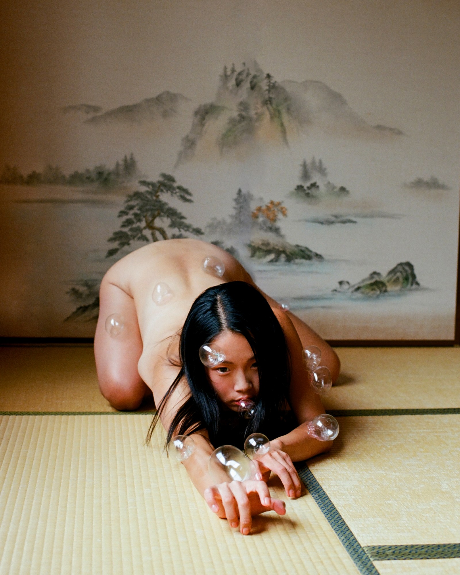 A nude woman chasing bubbles on a tatami floor, with a painted vista of pine trees and a cloudy mountain behind her