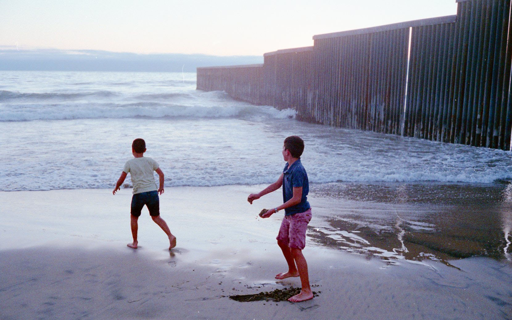 Two young boys play in the sand on the beach as the sun begins to set. Next to a wall creating a border between the United States and Mexico.”