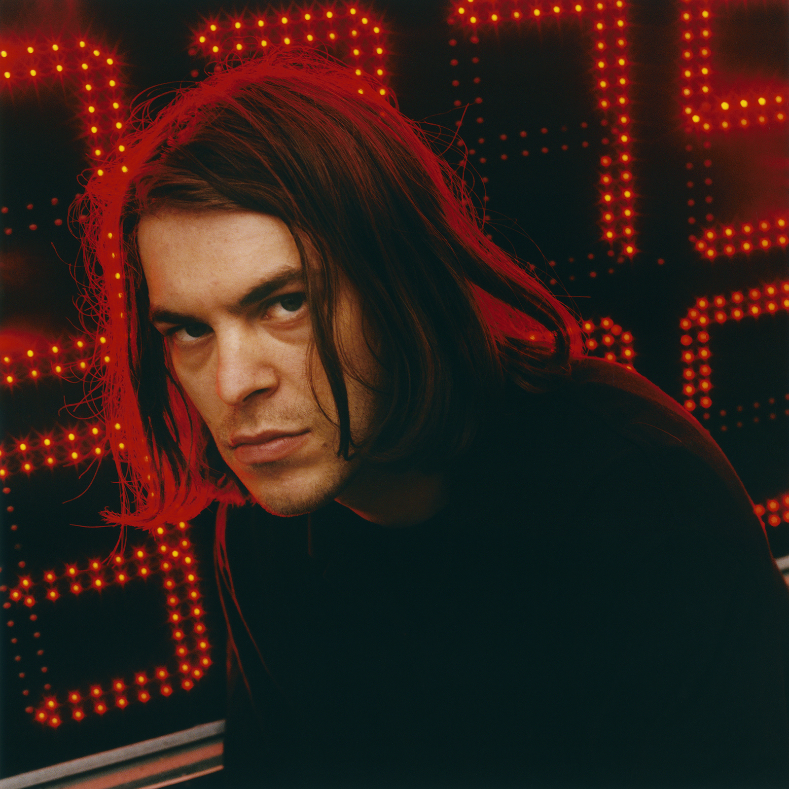 a man with shoulder length hair sits in front of a neon red sign