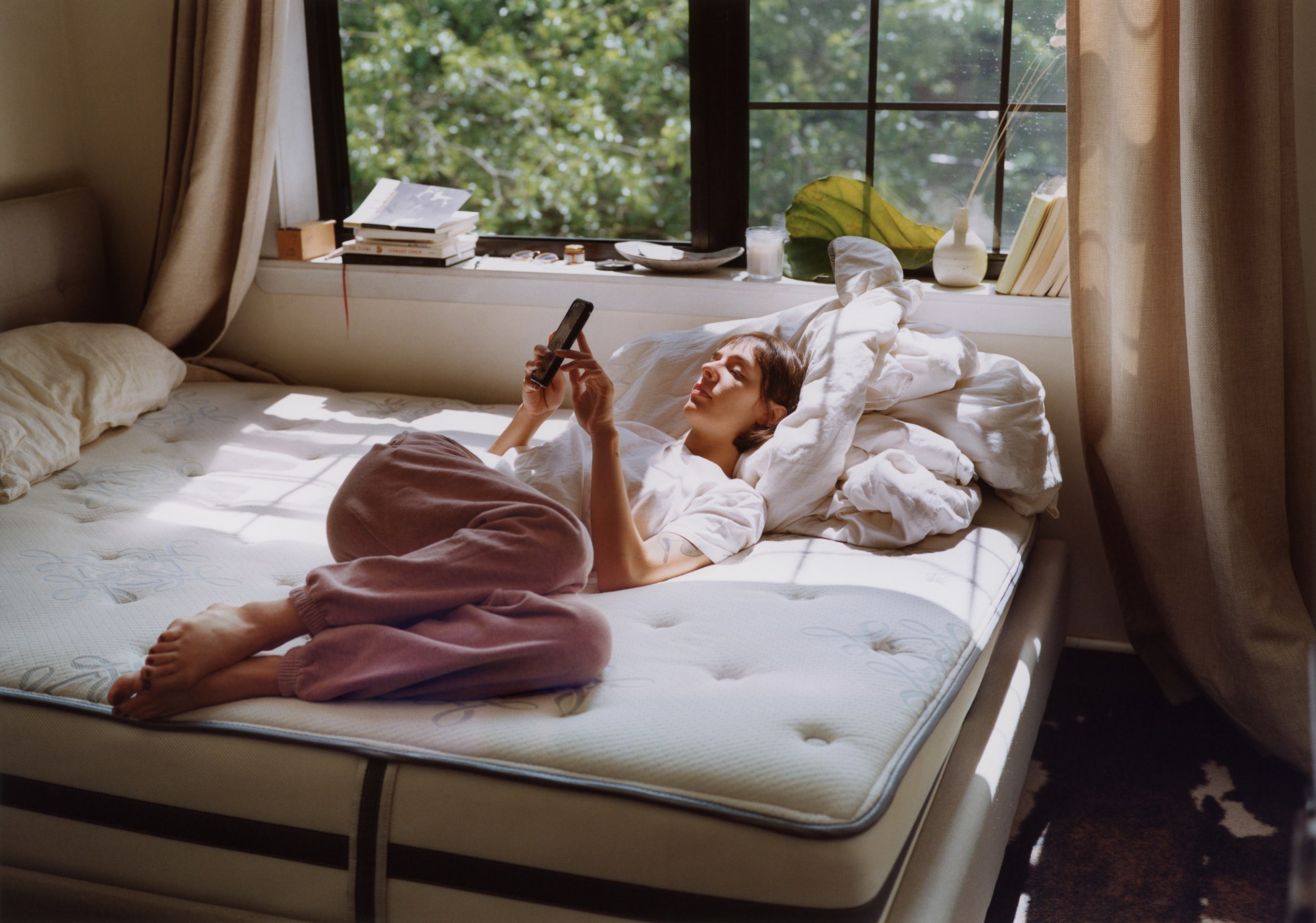 A woman lying on a bed in front of a window, looking at a phone