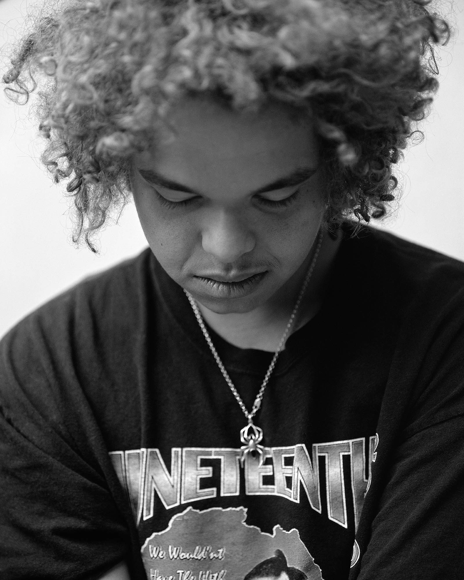 a man wearing a spider necklace and graphic print tee stares at the floor