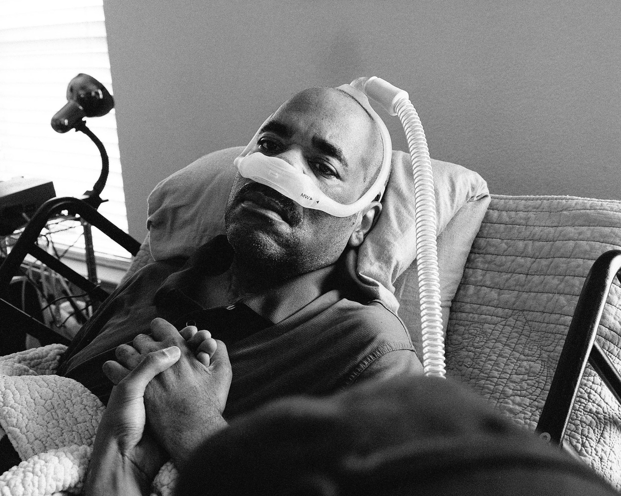 a man lies in bed holding someone's hand with a ventilator over his face