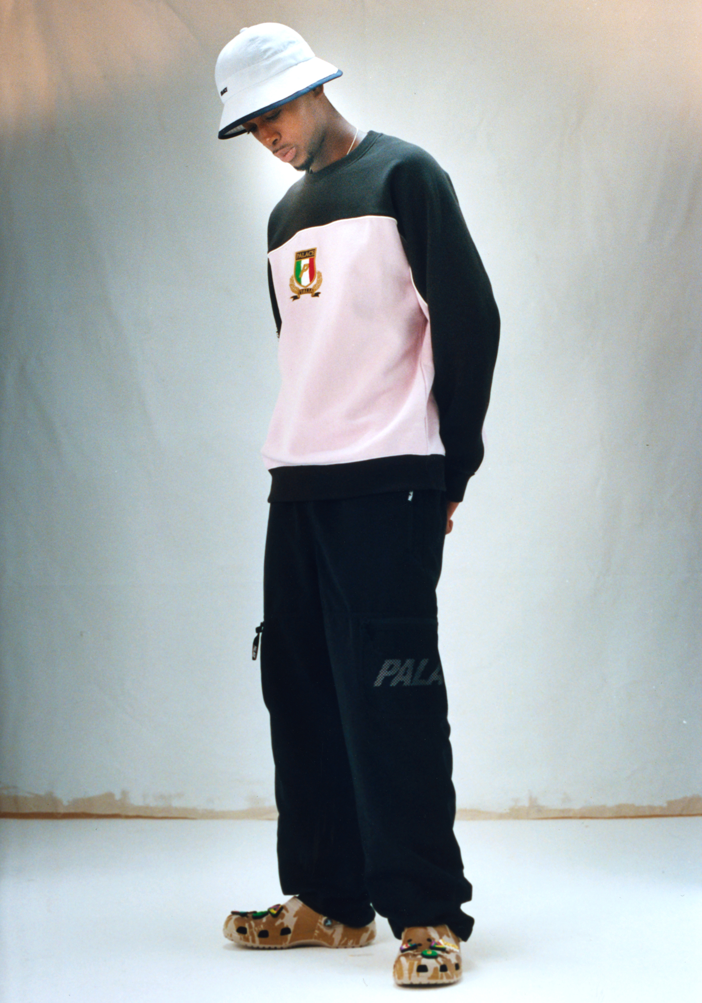 Model and skater Kyle Wilson looks to his feet wearing a pink Palace sweater, black sweatpants and camouflage Croc shoes