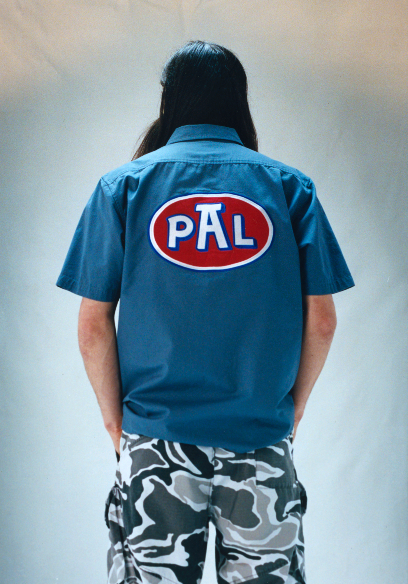 Skater and model Charlie Birch wears a gas station worker-style shirt and grey camouflage pants, looking away from the camera