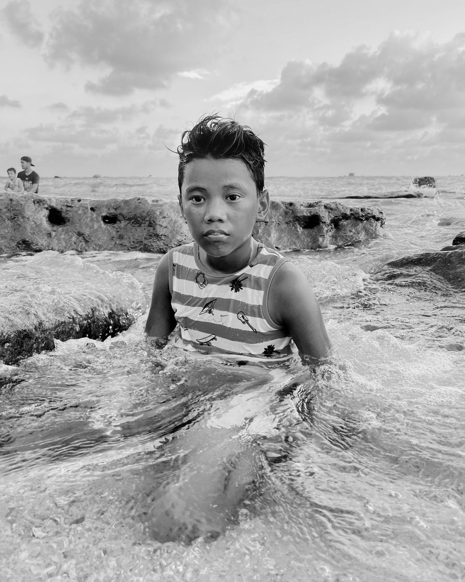 a young boy sits in the shallows, looking to camera as the water laps around him