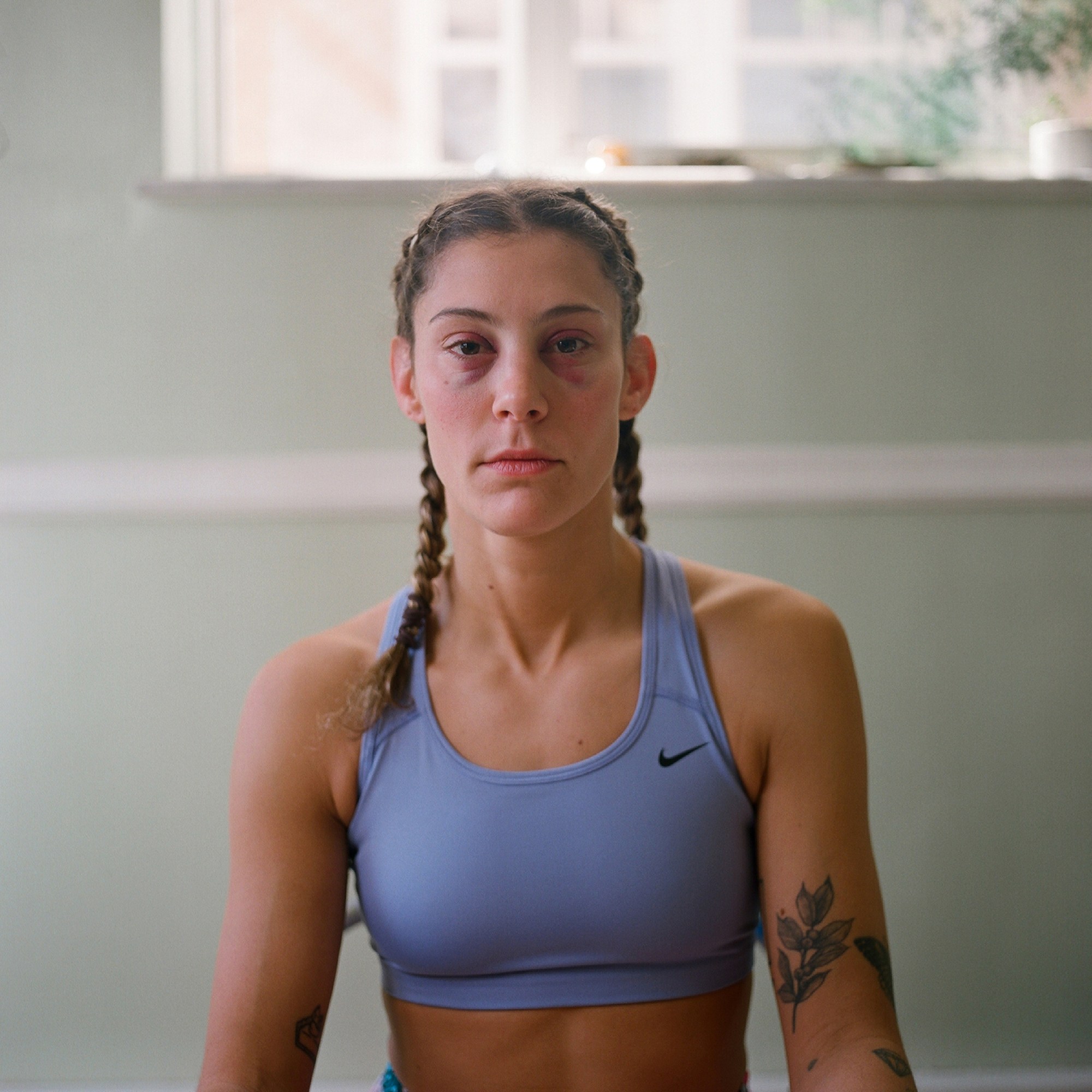 a tattooed woman wearing a lavender nike sports bra, her hair in two braids, with bruising around her eyes