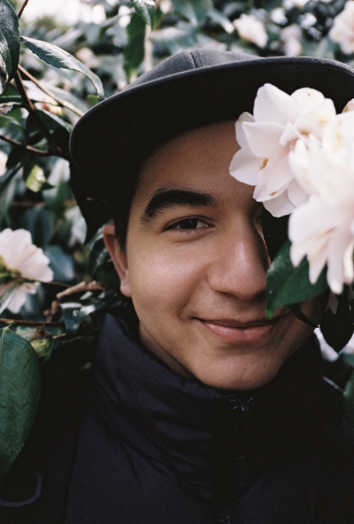 a man wearing a black cap smiles from behind a pink flower