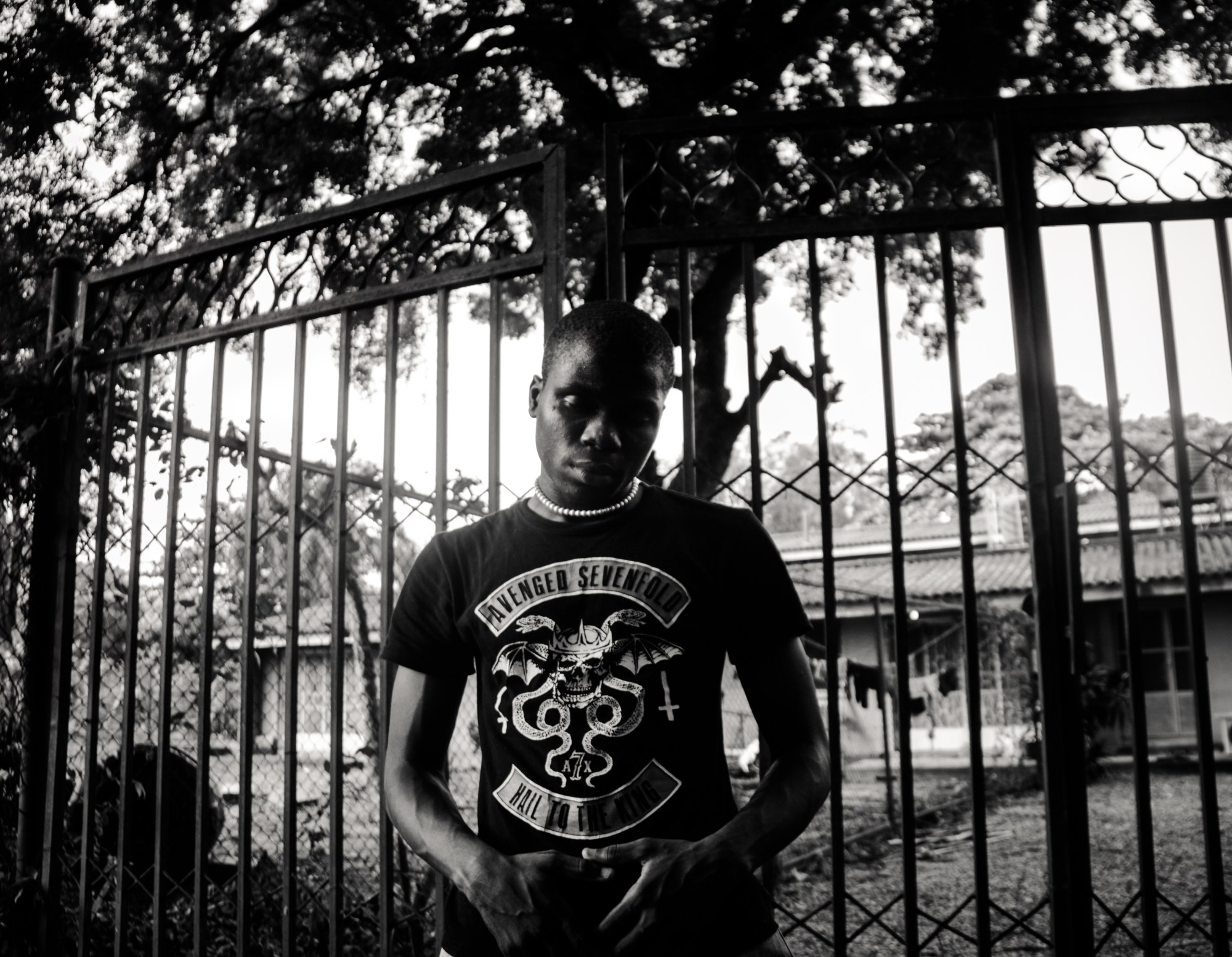 a black-and-white shot of a man standing in front of a gate in an Avenged Sevenfold T-shirt