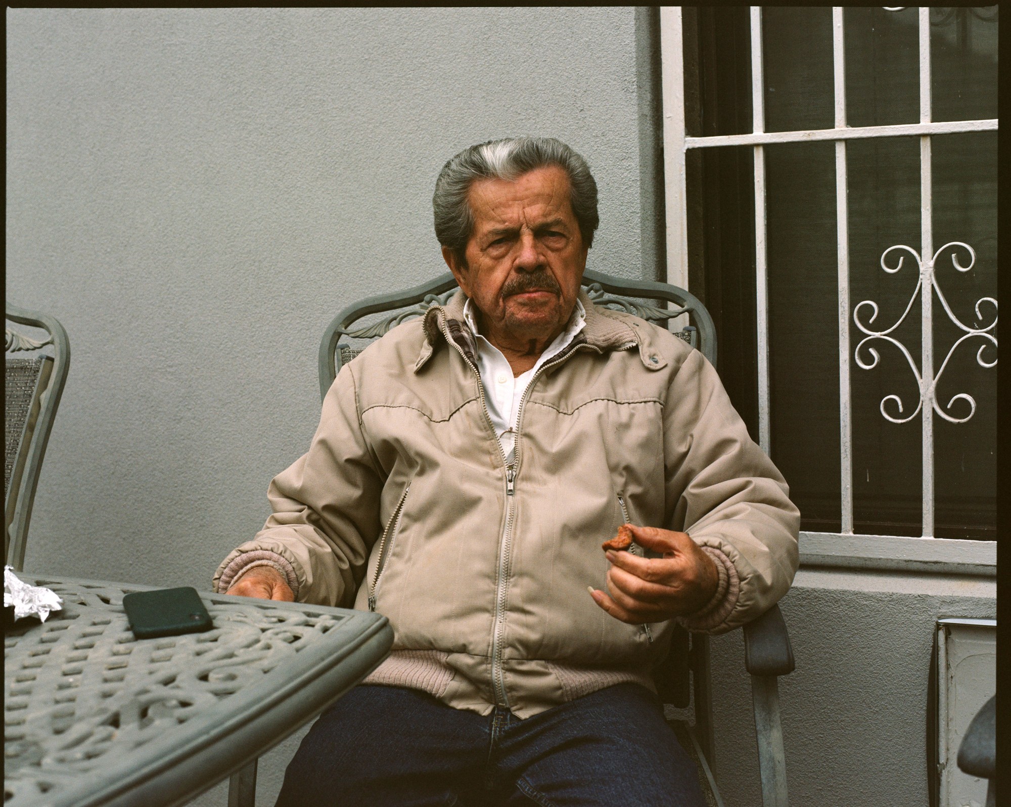 an old man in a beige jacket with silver hair, sitting outside at a table