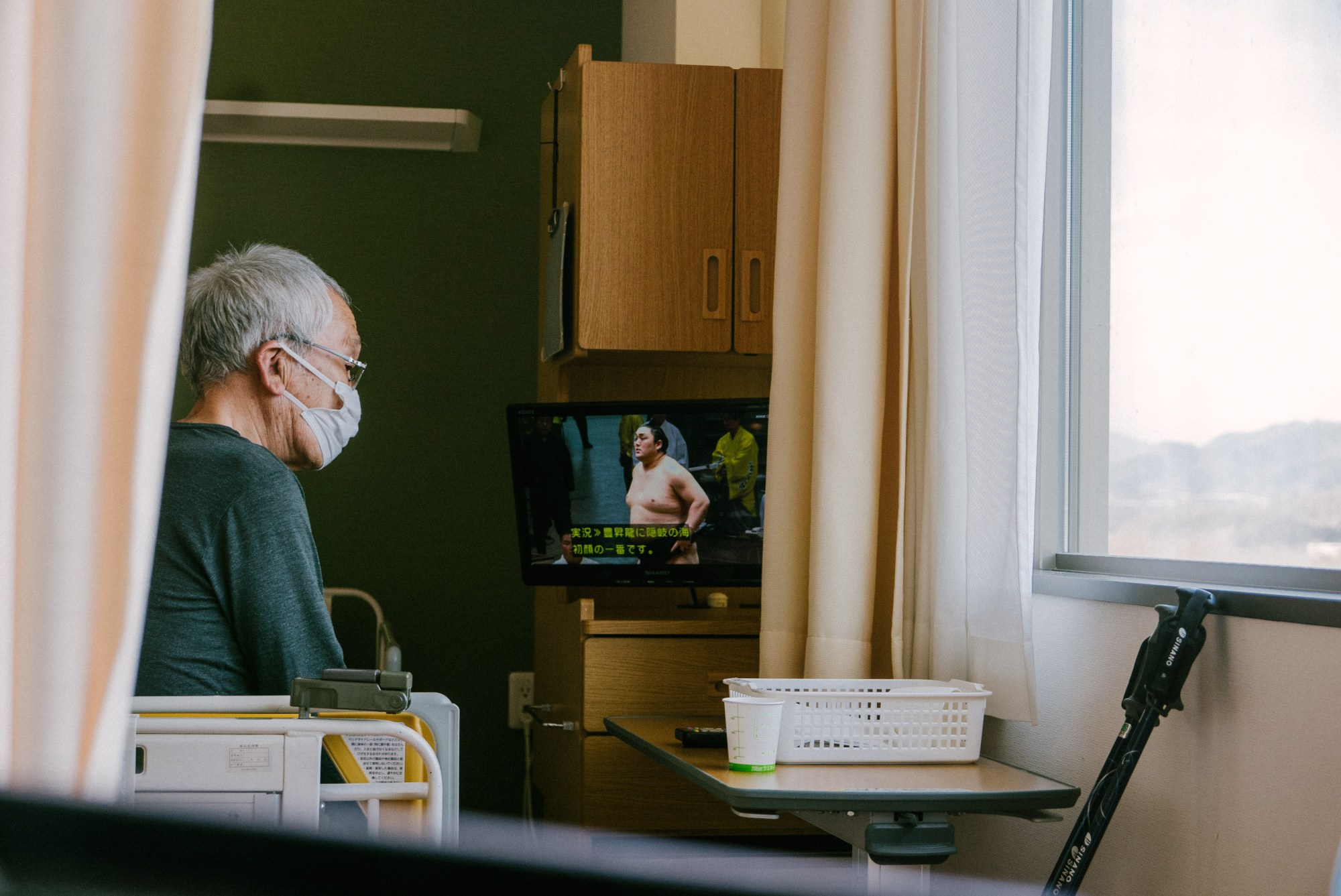 an old japanese man wearing a mask sits on a hospital bed watching sumo wrestling on TV