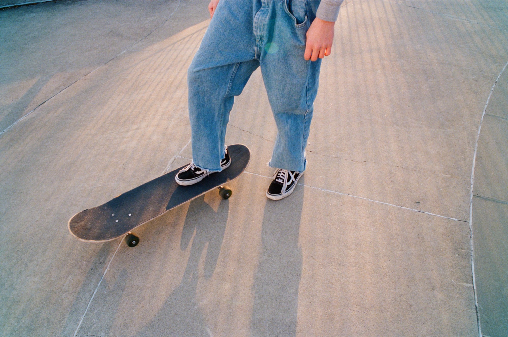 a person in oversized denim overalls stands with their foot on a skateboard in the sunlight