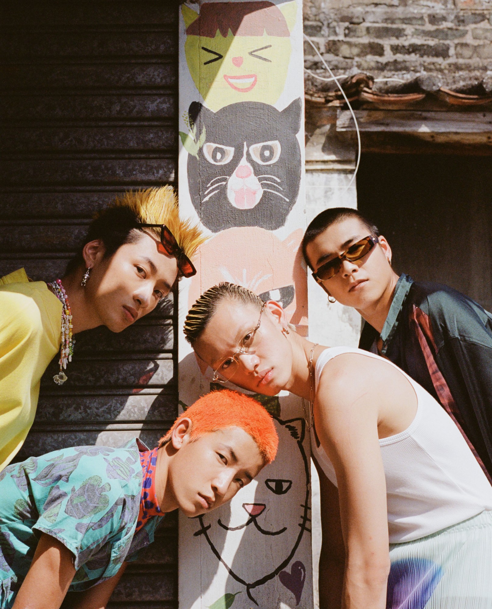 four young men with colourful hair lean into shot in front of a wall painted with a totem of cat heads