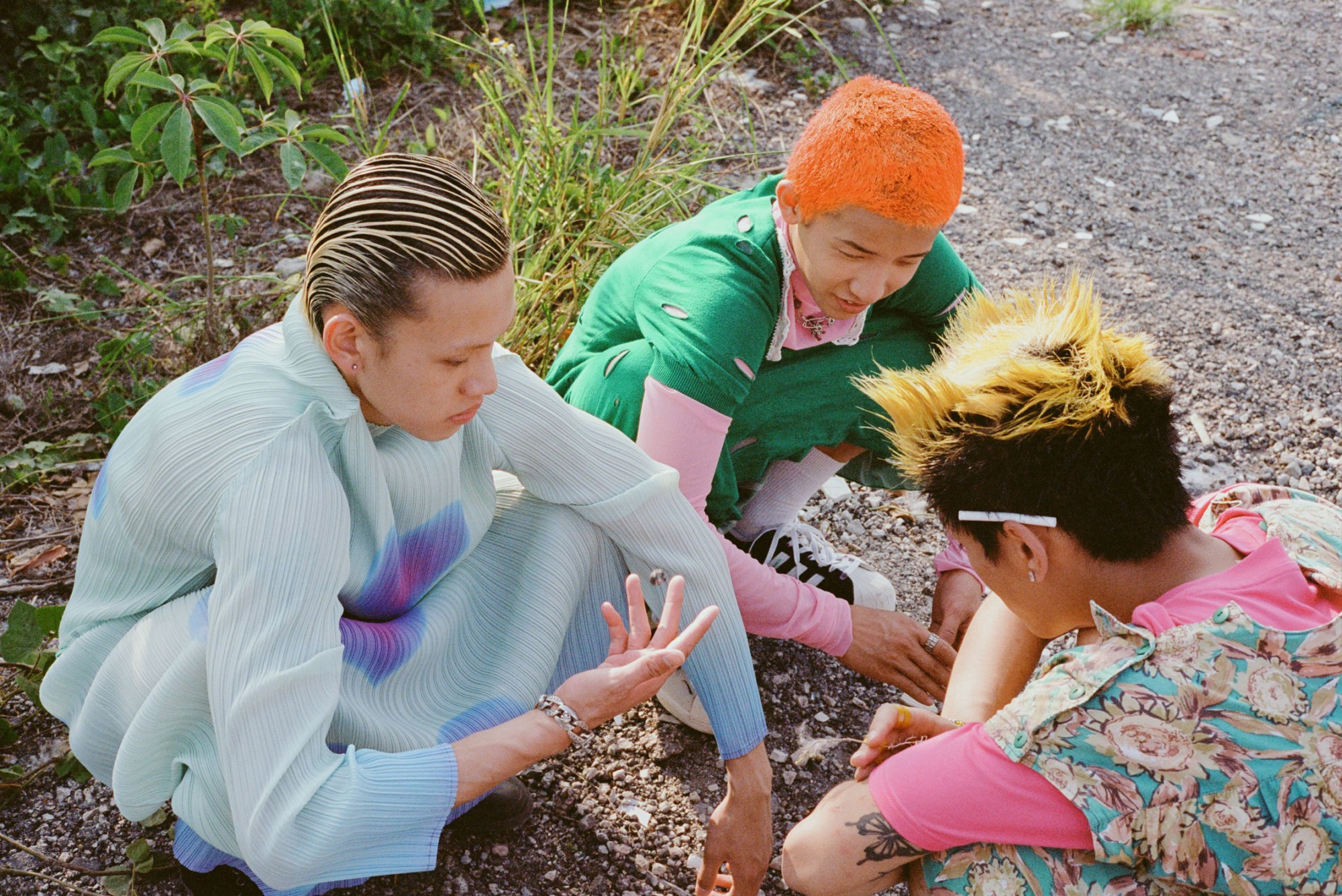 three young men with colourful hair wear bright dresses as they squat on a gravel floor, talking